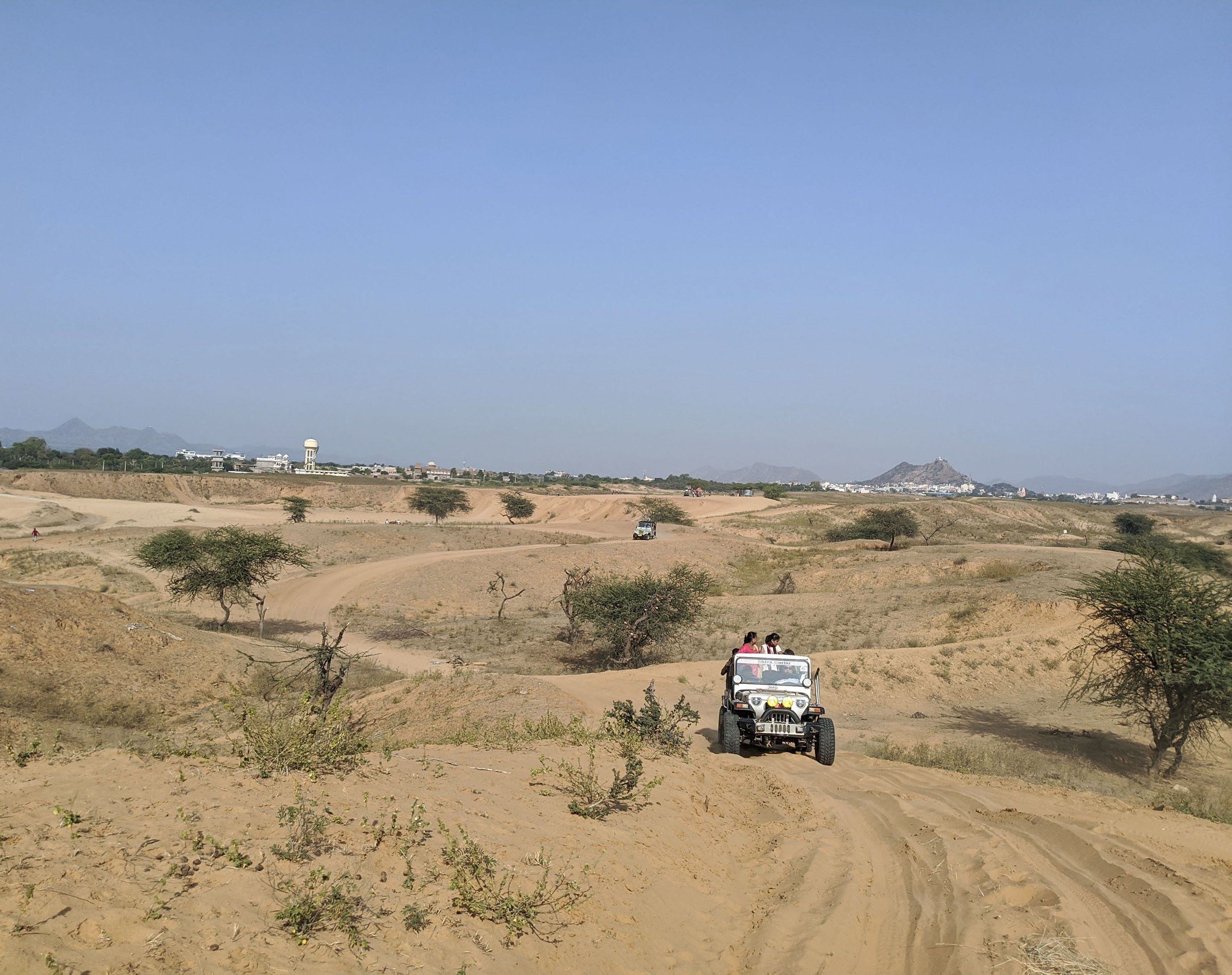 An off-road jeep safari navigating through sandy desert terrain near a town in Rajasthan, India, with scattered shrubs and distant hills under a clear blue sky.