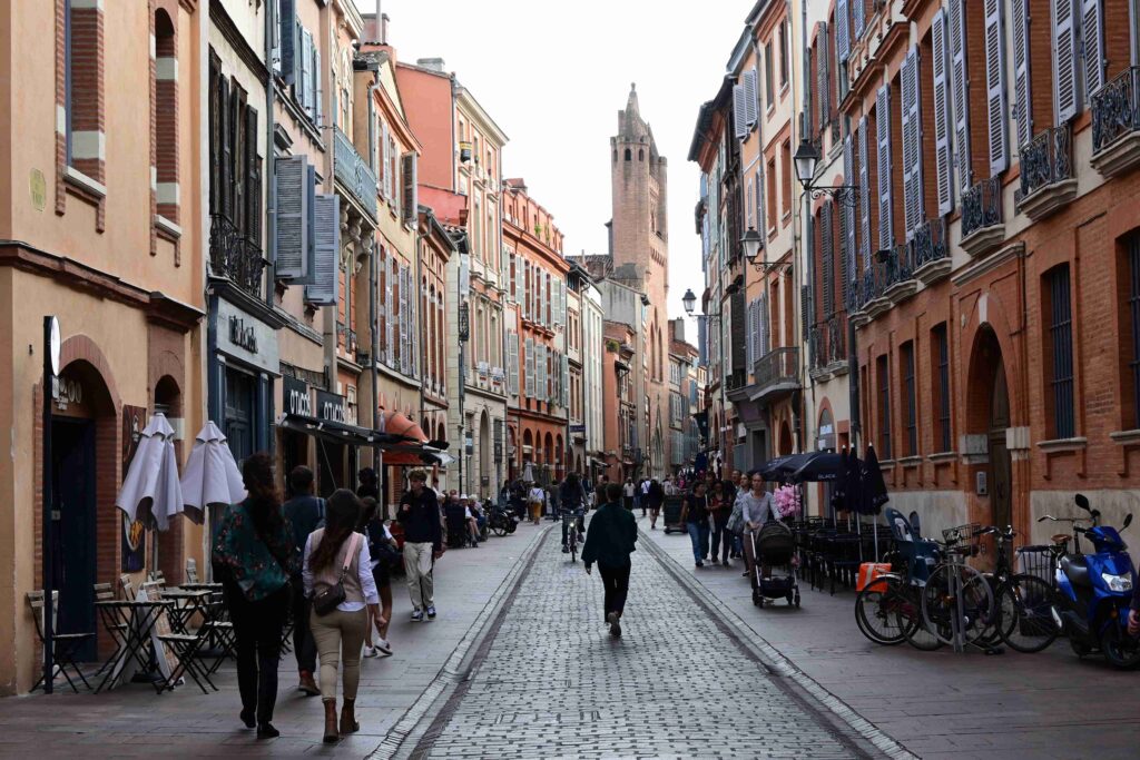 A charming cobblestone street in Toulouse, France, lined with historic red-brick and pastel-colored buildings featuring shuttered windows and wrought-iron balconies. People stroll along the lively pedestrian street, with outdoor cafés, shops, and bicycles adding to the vibrant atmosphere. In the background, the tall brick tower of a historic church rises above the rooftops.