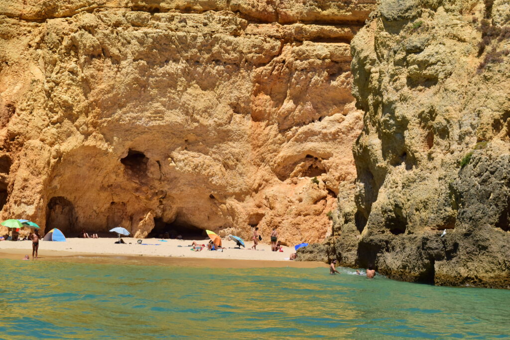 A beautiful sun kissed beach as seen from the water in Algarve, Portugal. There are tall sandstone cliffs in the backdrop and the beach has a tiny portion of sand. There are people with towels and umbrellas on the sand and swimmers in the water.