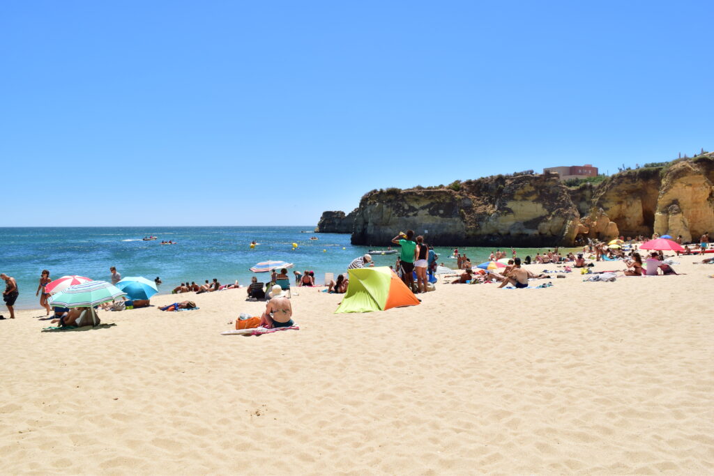 A gorgeous beach on a sunny day without a cloud in the sky. The picture shows many colorful towels and people sun bathing on the beach with clear blue, turquoise waters in the background. There are surfers and swimmers in the water. There is also a sandstone sea cliff the forms the boundary of the beach.