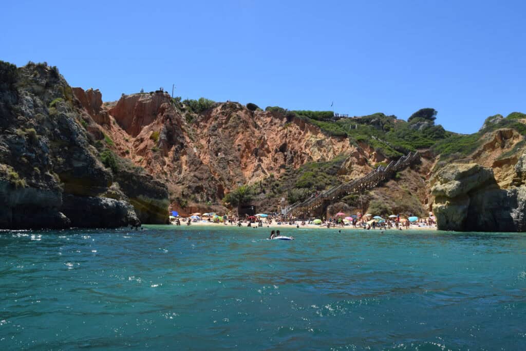 A gorgeous beach with a red sandstone sea cliff in the backdrop. There are stairs that lead down from the top of the cliff to the beach. There are many people lounging on the beach and in the water. 