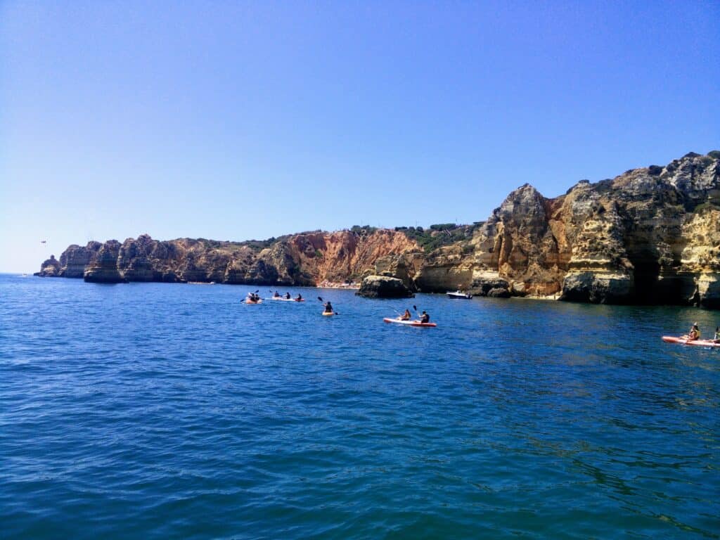 A picture of kayaks in the sea along red, coastal cliffs that line the right side of the image. The water is clear blue. 