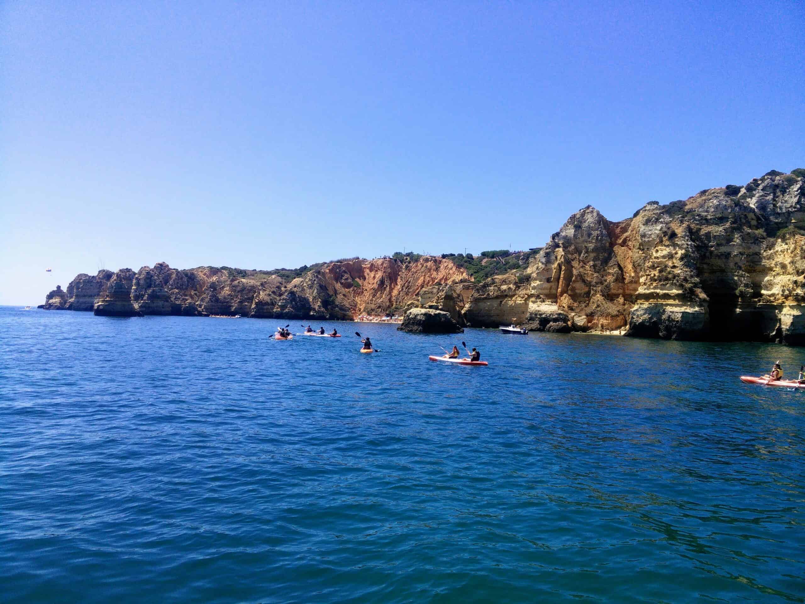 A picture of kayaks in the sea along red, coastal cliffs that line the right side of the image. The water is clear blue.