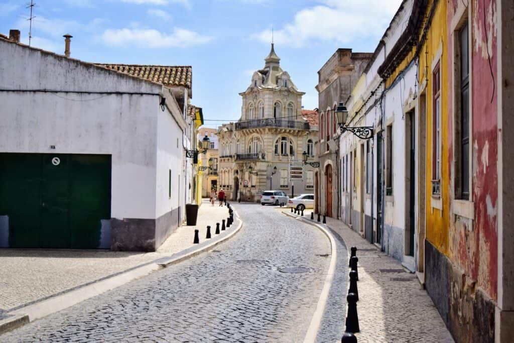 A picture of red, yellow, white buildings along a cobbled stone street in Faro, Portugal. There is an imposing medieval building in the background and a couple of cars of the street. 