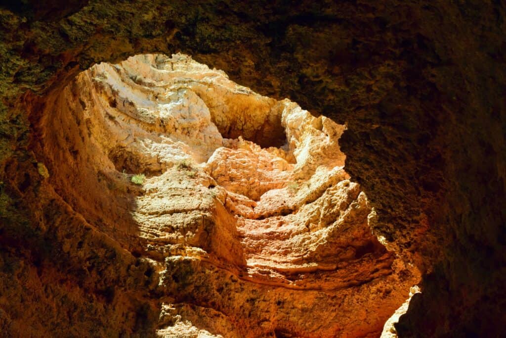 A picture of the limestone cliffs in Algarve, Portugal. The picture is a zoomed in shot of a hole in the cliff. The light peaks in through the hole creating a red, yellow and black contrast within the rocky surface. 