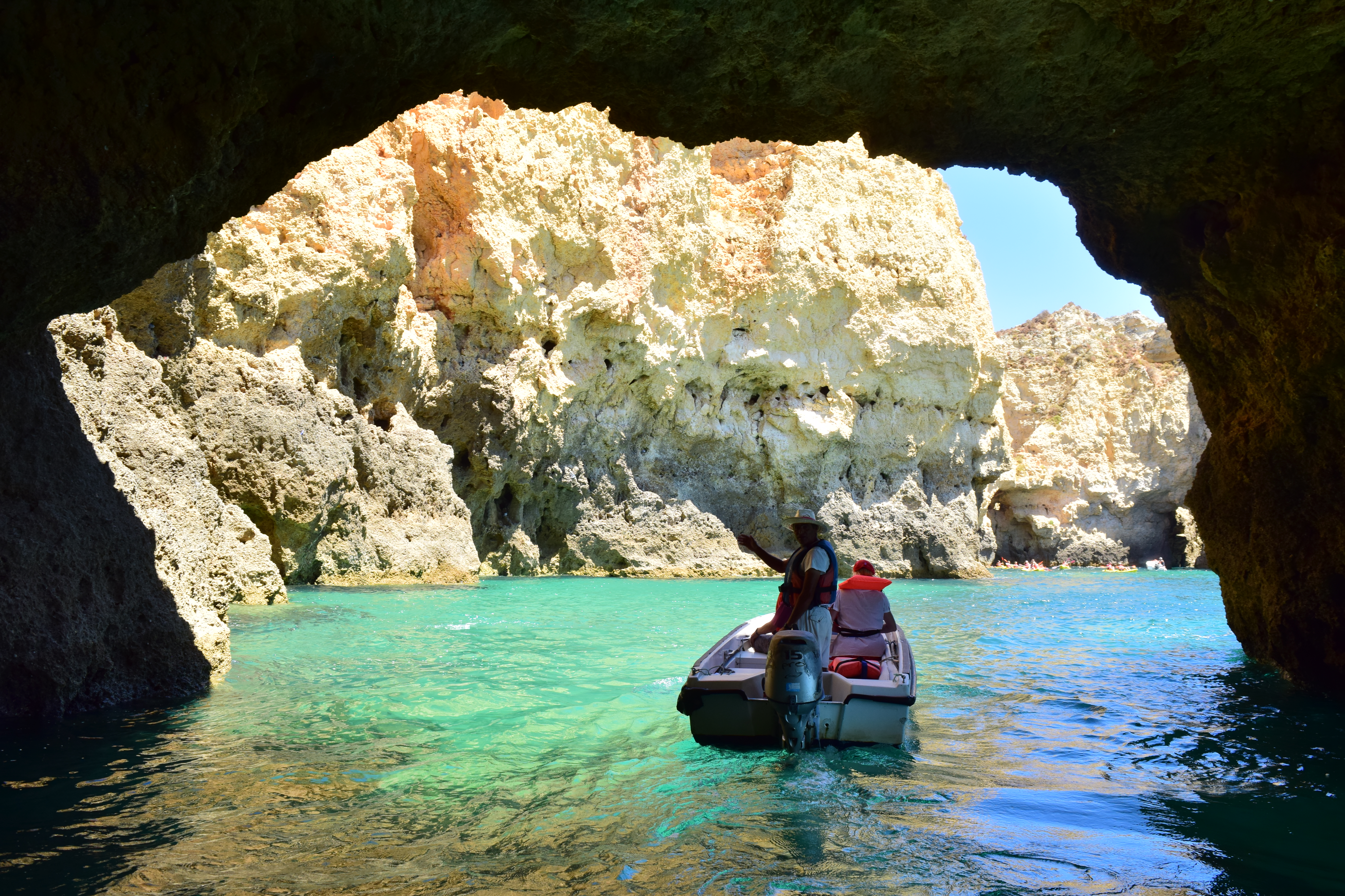 The picture has been taken at the Benagil caves in Algarve, Portugal. There is a boat in the picture with 3 on board inside a sea cave. You can see the clear, turquoise water and limestone sea cliffs in the background. 