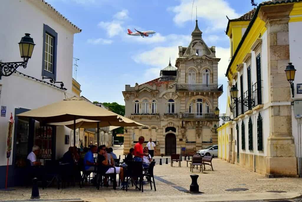 A picture of a medieval two floor building with a little square in the foreground. There are two other buildings along each side of the picture and are yellow and white. There are people at a restaurant in the square. A plane can be seen in the background in the blue sky behind the building. 