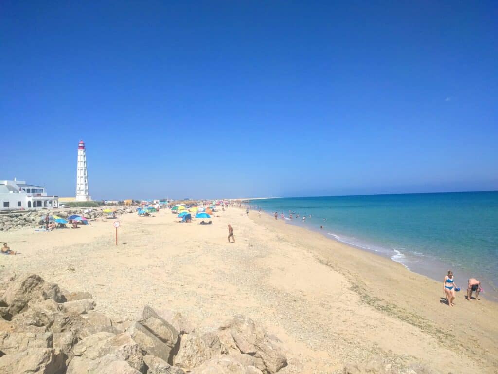 A picture of a lengthy beach with white sand and clear, turquoise waters. The picture is in Faro, Portugal and you can see a lighthouse in the background. There are people lounging along the entire length of the beach.