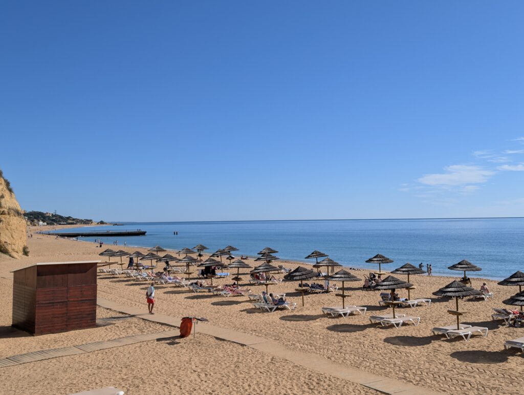 A gorgeous beach in Albufeira, Algarve, South Portugal. The picture shows sun beds and umbrellas lining the beach with blue waters and a clear blue sky in the background. 