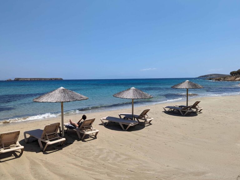 Golden Beach in Paros, Greece with straw sun umbrellas and empty lounge chairs on soft sand, overlooking calm turquoise water and a distant rocky islet on a clear sunny day.