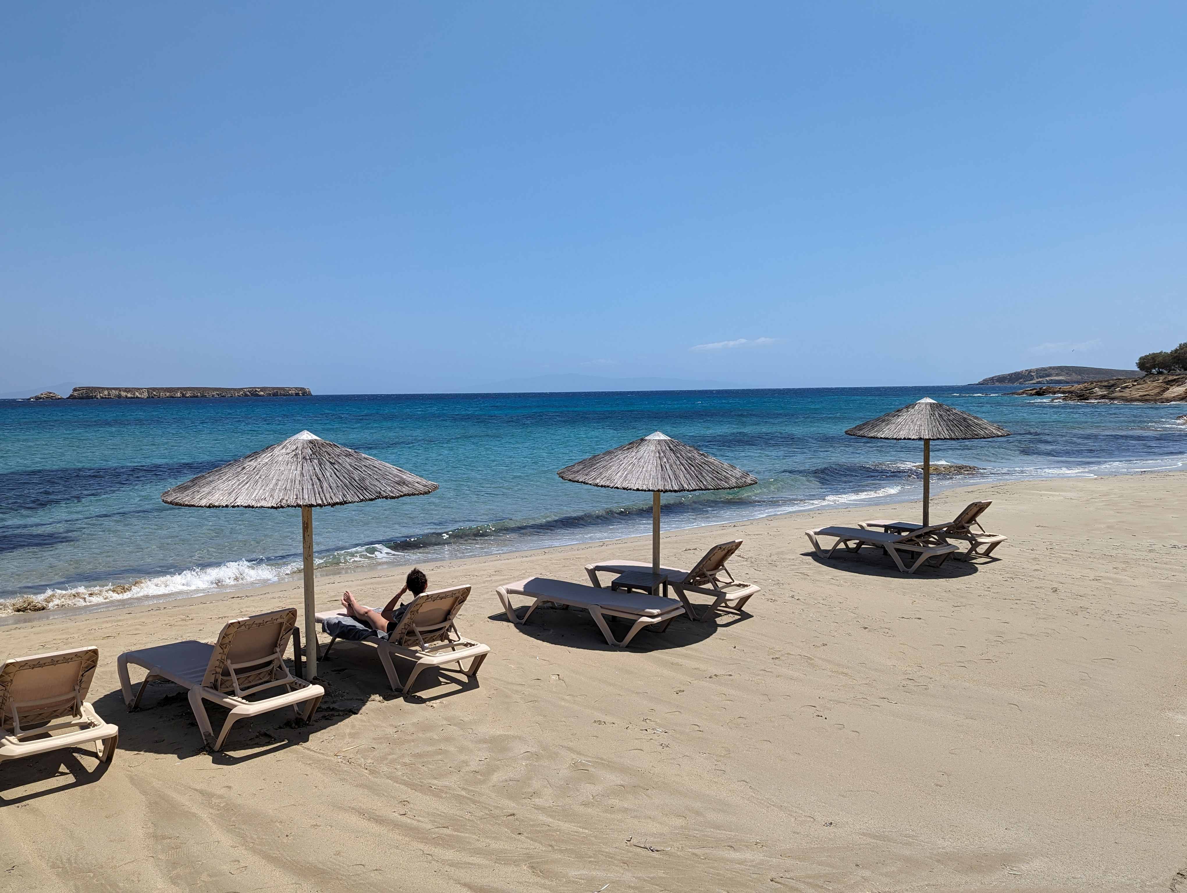 Golden Beach in Paros, Greece with straw sun umbrellas and empty lounge chairs on soft sand, overlooking calm turquoise water and a distant rocky islet on a clear sunny day.