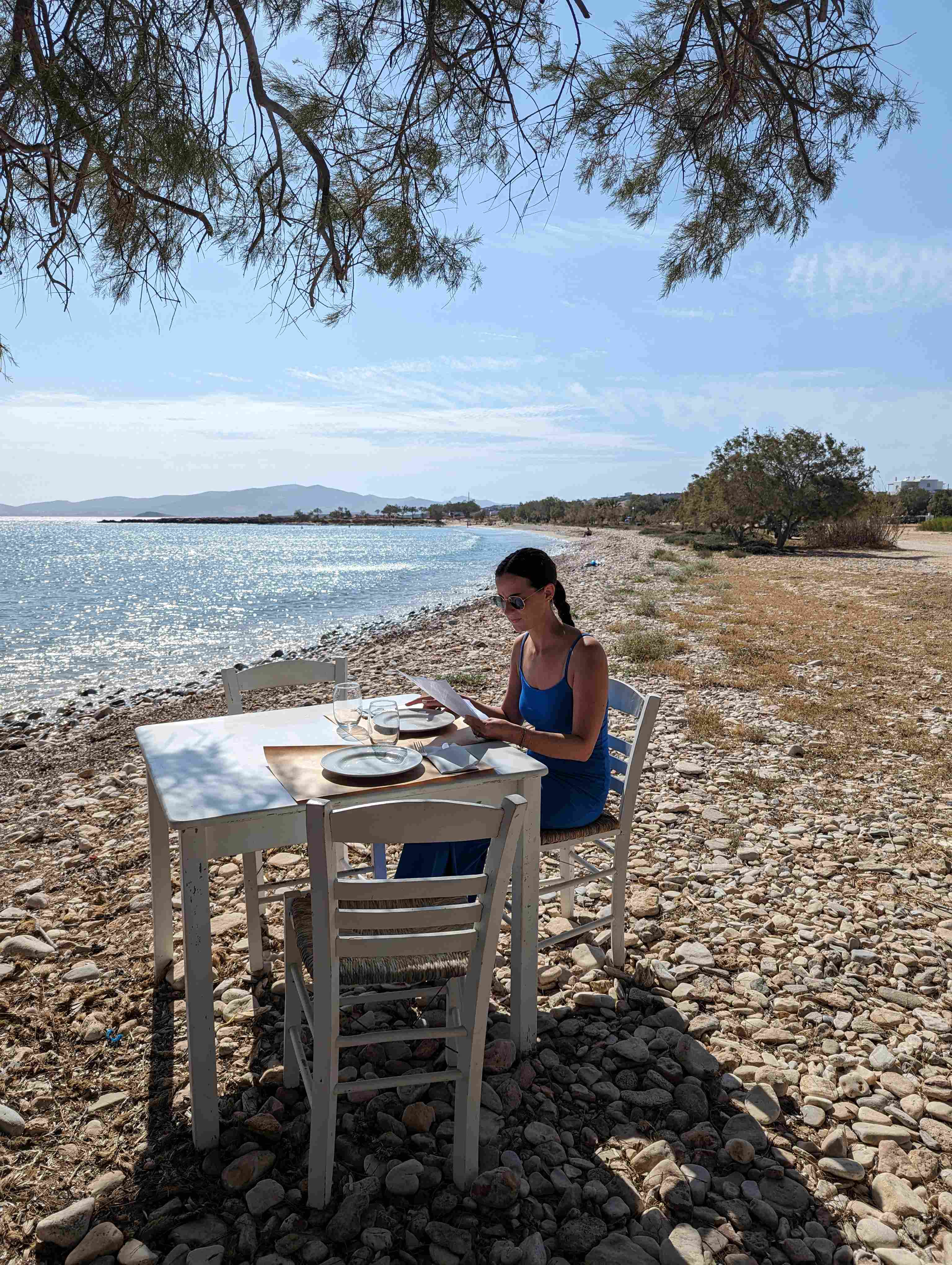 Woman dining alone at white table on pebble beach under tree branches with sparkling sea in the background in Aliki on Paros, Greece.