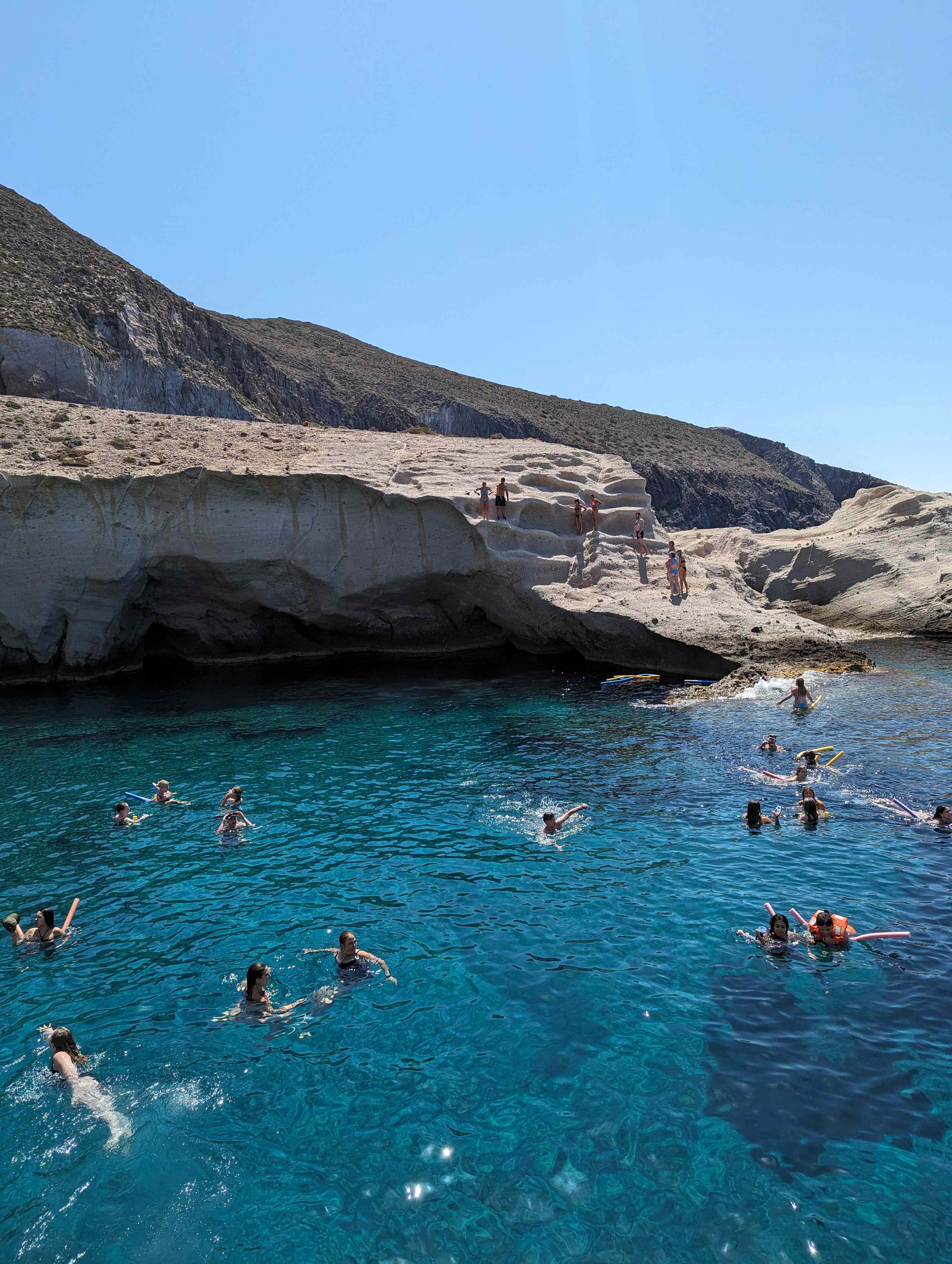 People swimming in crystal-clear turquoise water near white rocky cliffs on a sunny day during a boat tour around Paros, Greece