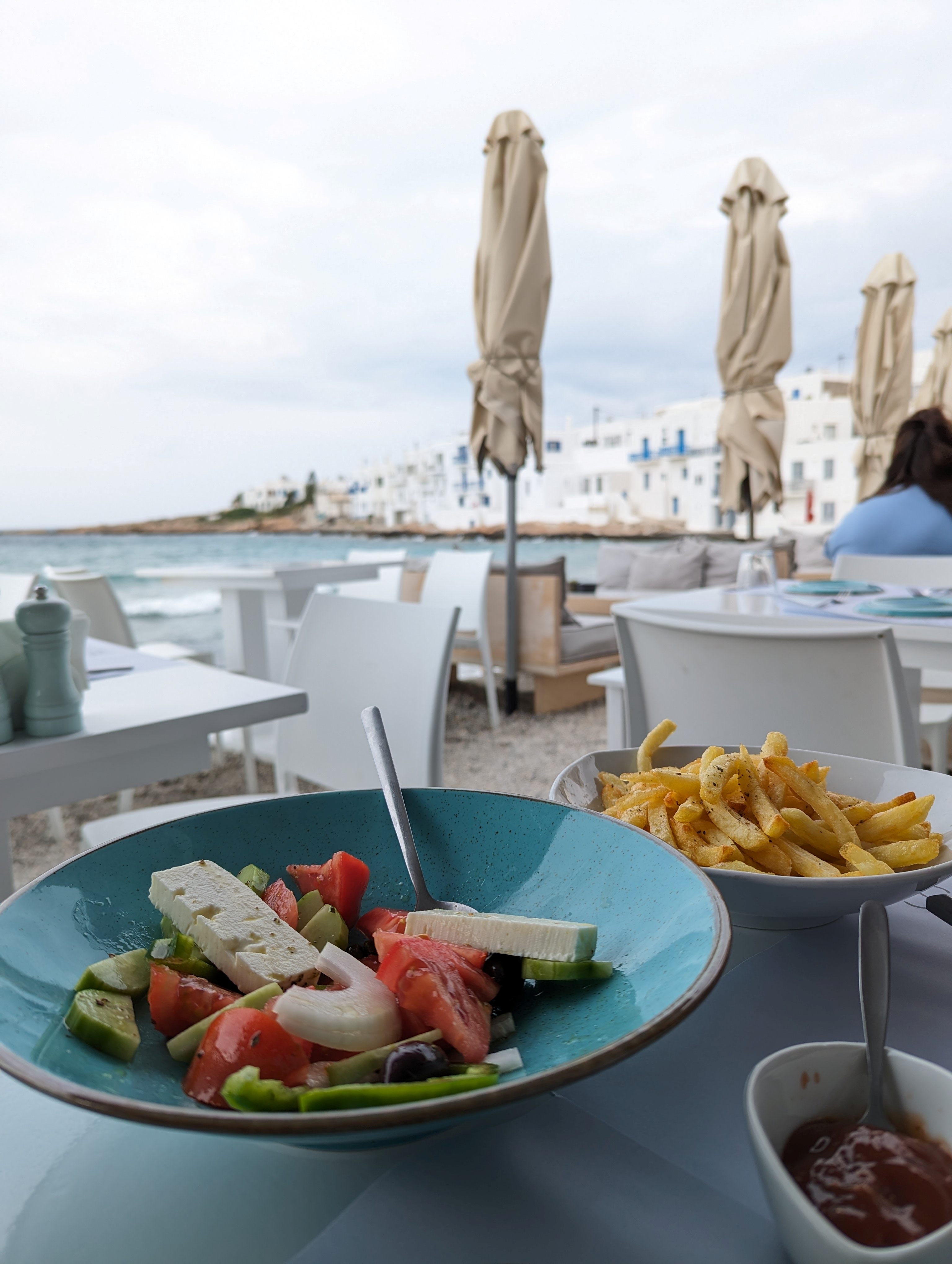 Mediterranean waterfront dining with Greek salad and french fries on table overlooking white buildings and blue sea.