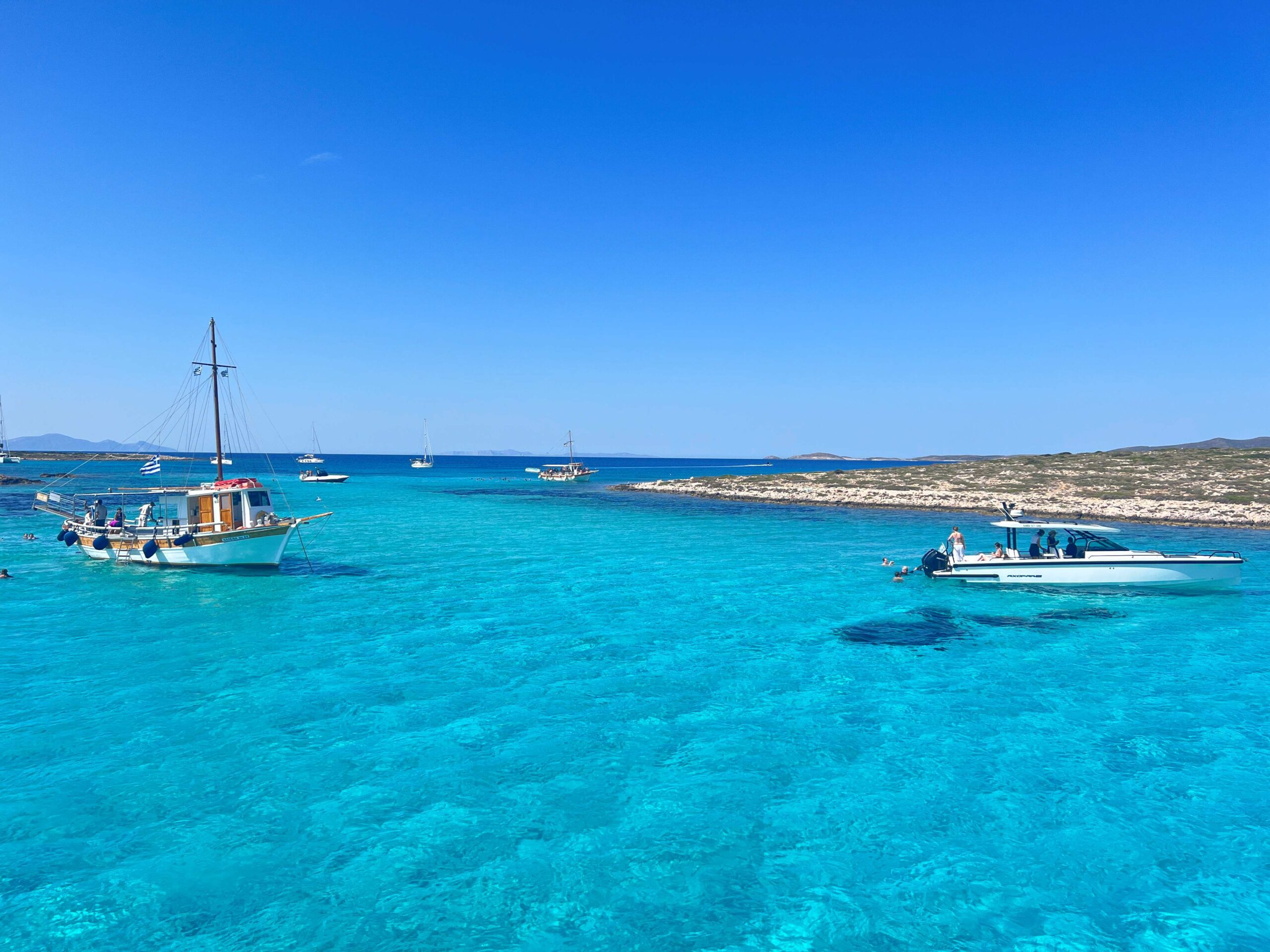Crystal-clear turquoise waters surround several boats anchored near a rocky island coastline under a bright blue sky. People are swimming and relaxing on the boats, enjoying a sunny day in the Aegean Sea near Paros, Greece.