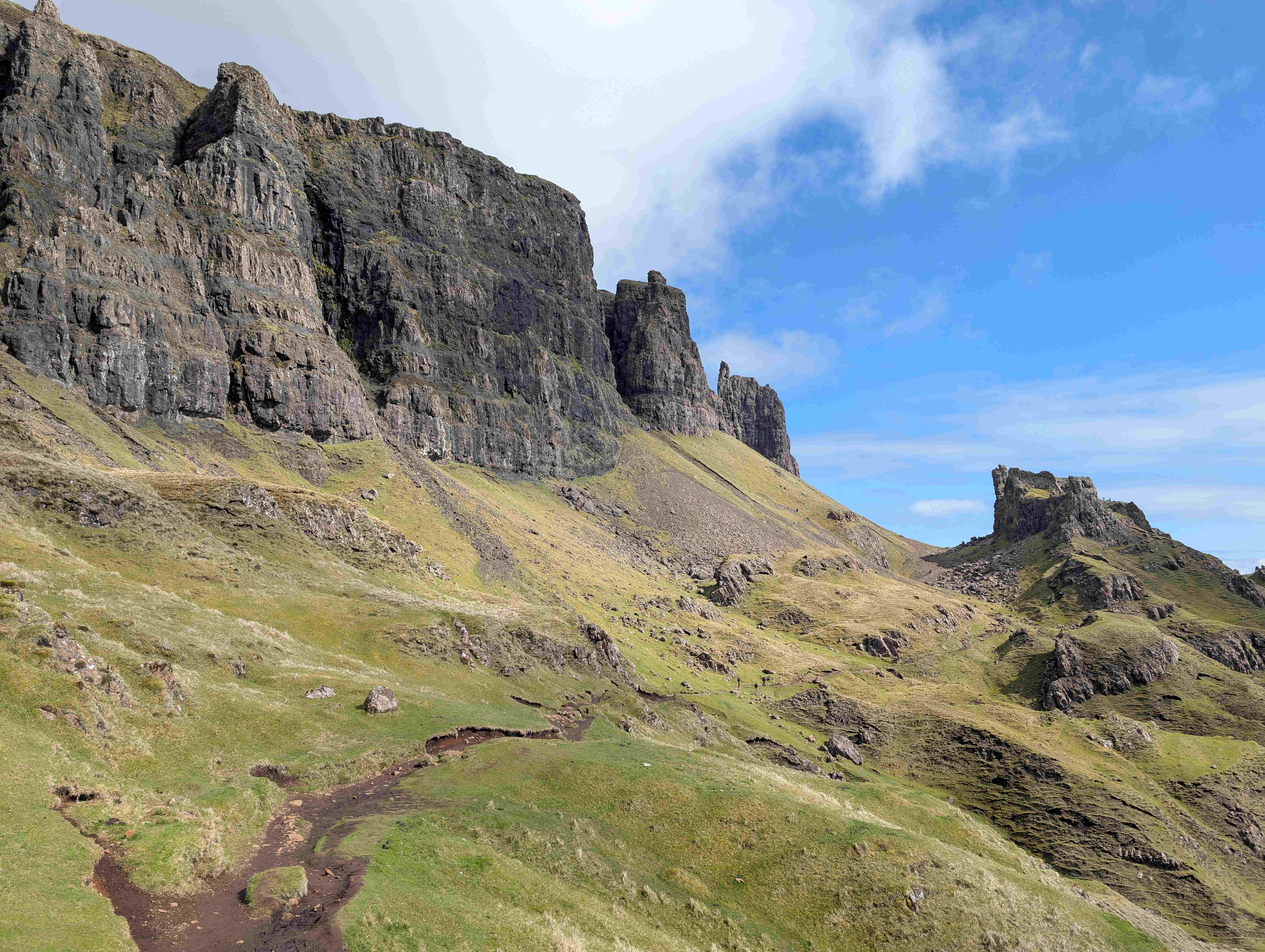 Scenic view of the Quiraing rock formations on the Isle of Skye, Scotland, featuring dramatic cliffs, jagged peaks, and a rugged grassy hillside under a bright blue sky with scattered clouds. A dirt hiking path winds through the foreground.