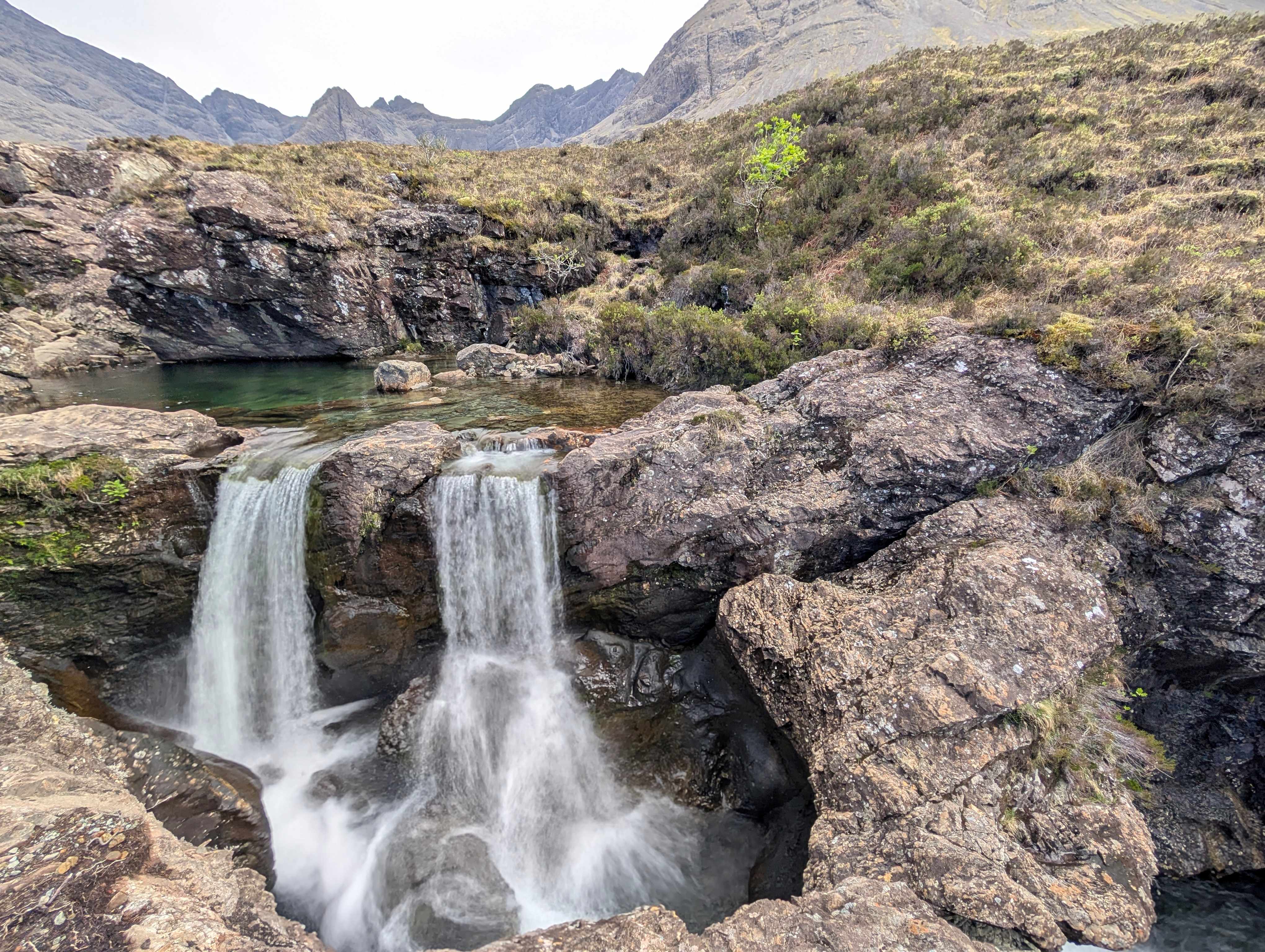 A small cascading double waterfall flows into a clear turquoise pool surrounded by rugged rocks at the Fairy Pools on the Isle of Skye, Scotland. In the background, the jagged peaks of the Cuillin Mountains rise under an overcast sky.


