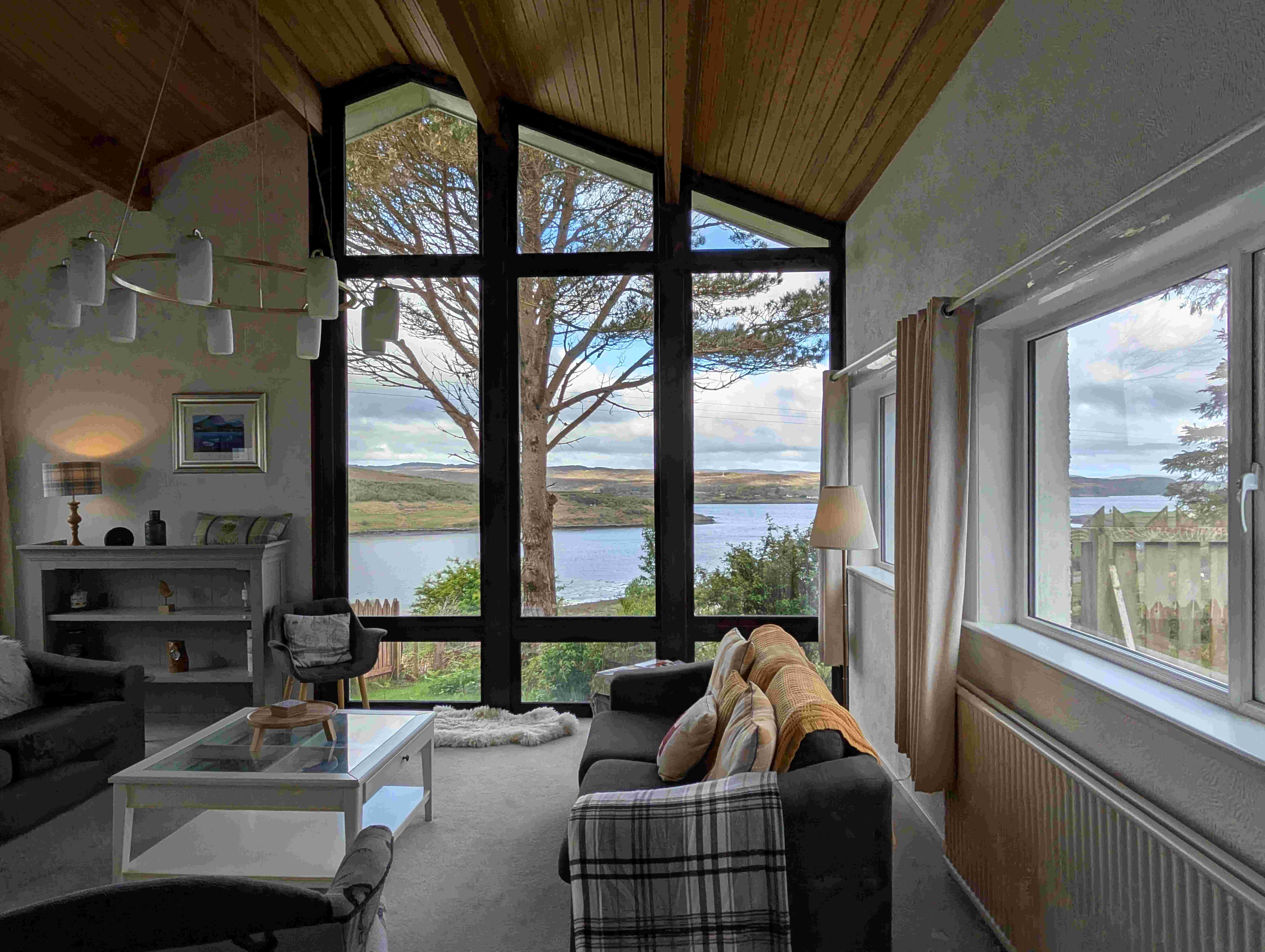 Cozy living room interior at Loch Eyre House on the Isle of Skye, featuring large floor-to-ceiling windows with stunning views of the loch and rolling hills beyond. The space includes a dark grey sofa with plaid and mustard throw pillows, a modern white coffee table, and a mix of contemporary and rustic decor. 