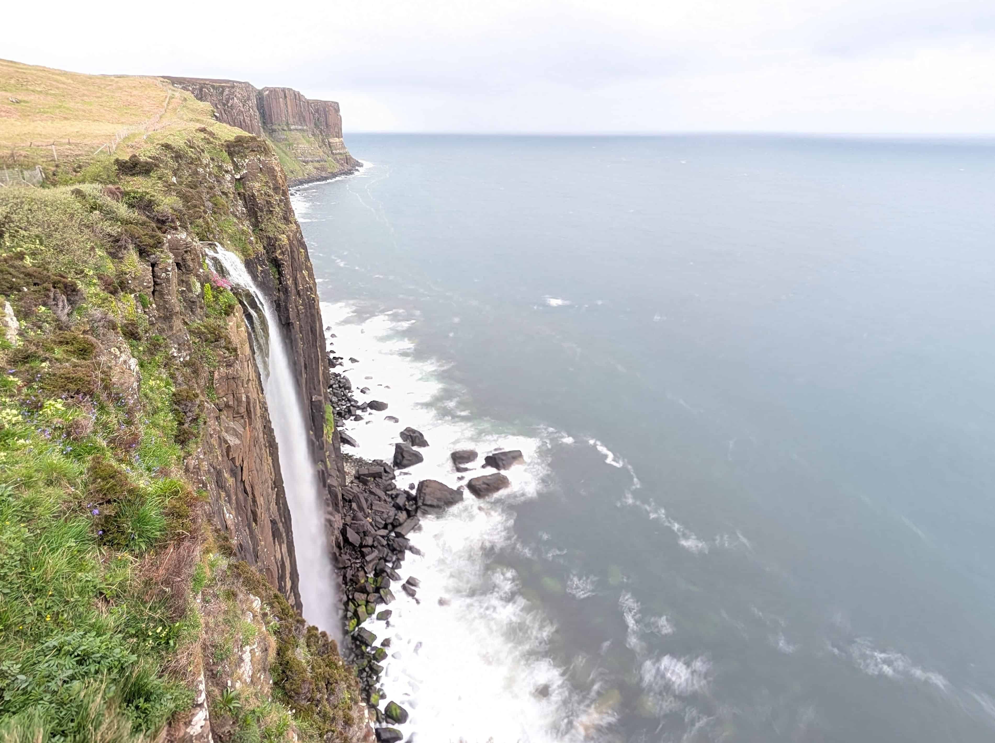 A dramatic view of Mealt Falls on the Isle of Skye, Scotland, where a powerful waterfall plunges off a rugged cliff directly into the ocean below. The surrounding sea cliffs stretch into the misty distance, and the Atlantic waves crash against dark rocks at the base of the cliff, creating a striking contrast with the green vegetation above.