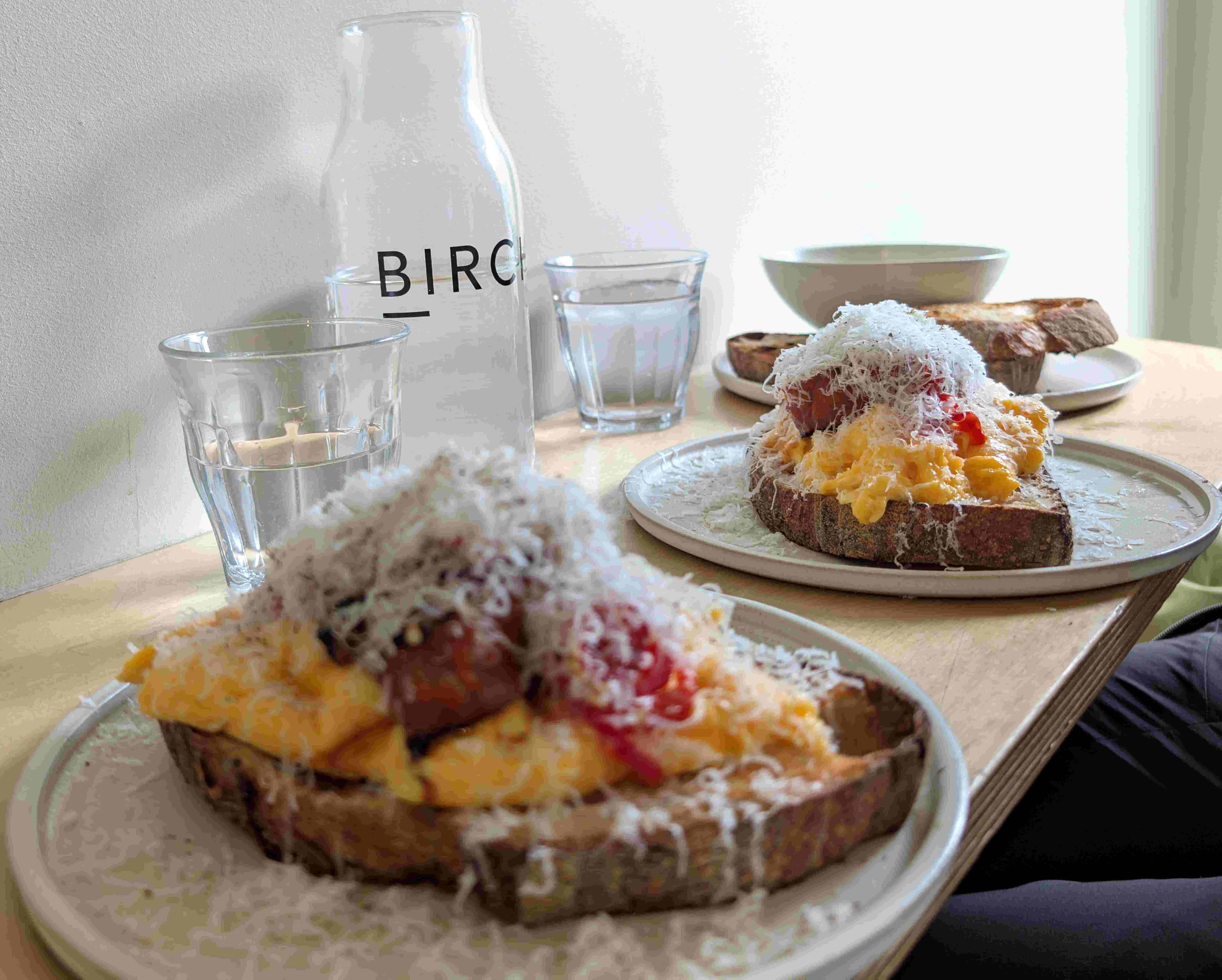 Two slices of dark bread topped with scrambled eggs, jam, and grated cheese on ceramic plates, with glasses and a water bottle labeled 'BIRCH' on a wooden table.
