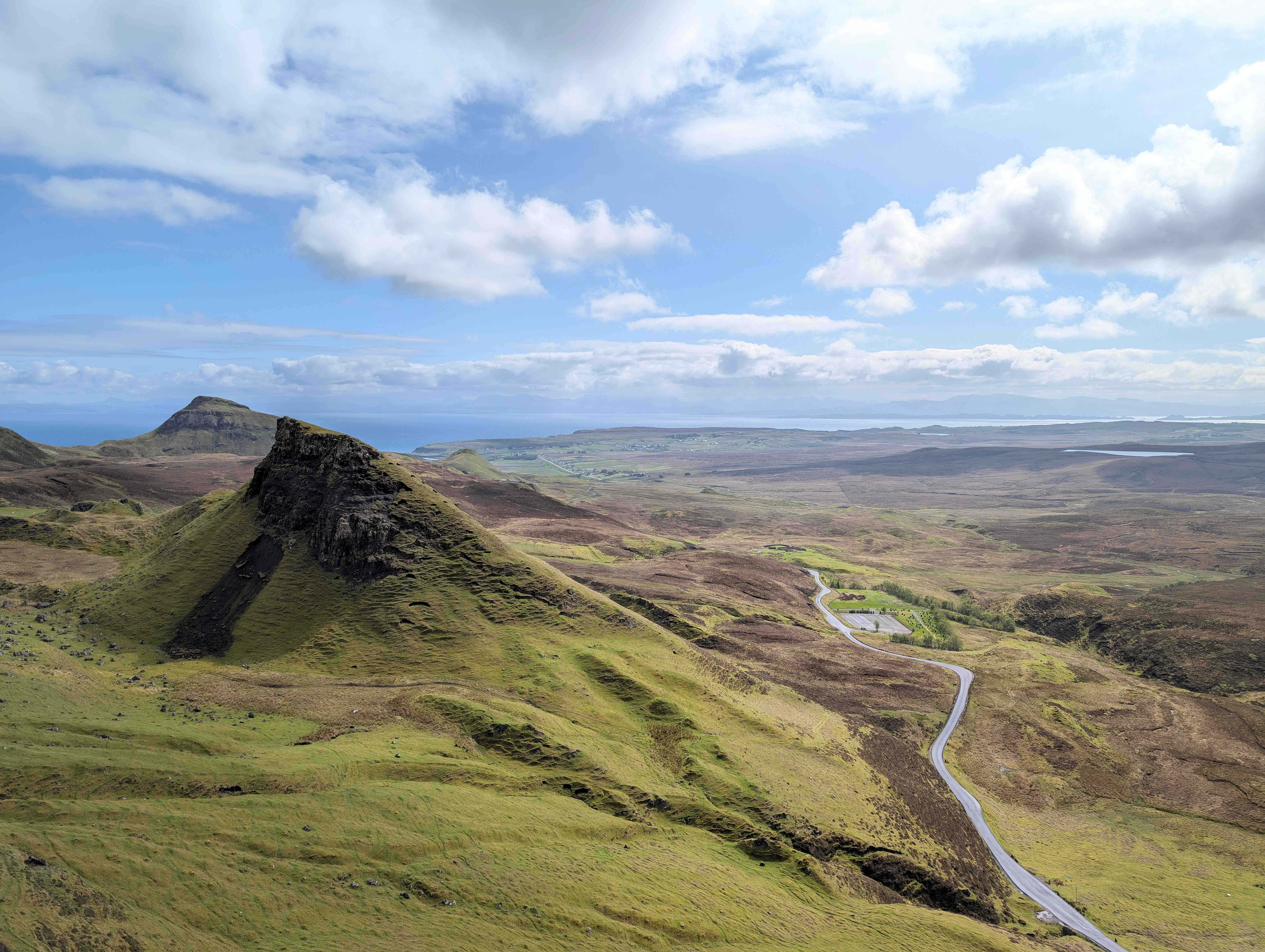Panoramic view of the Quiraing landslip on the Isle of Skye in Scotland, showcasing dramatic green hills, a winding road, and a partly cloudy sky stretching over a vast, rugged landscape.