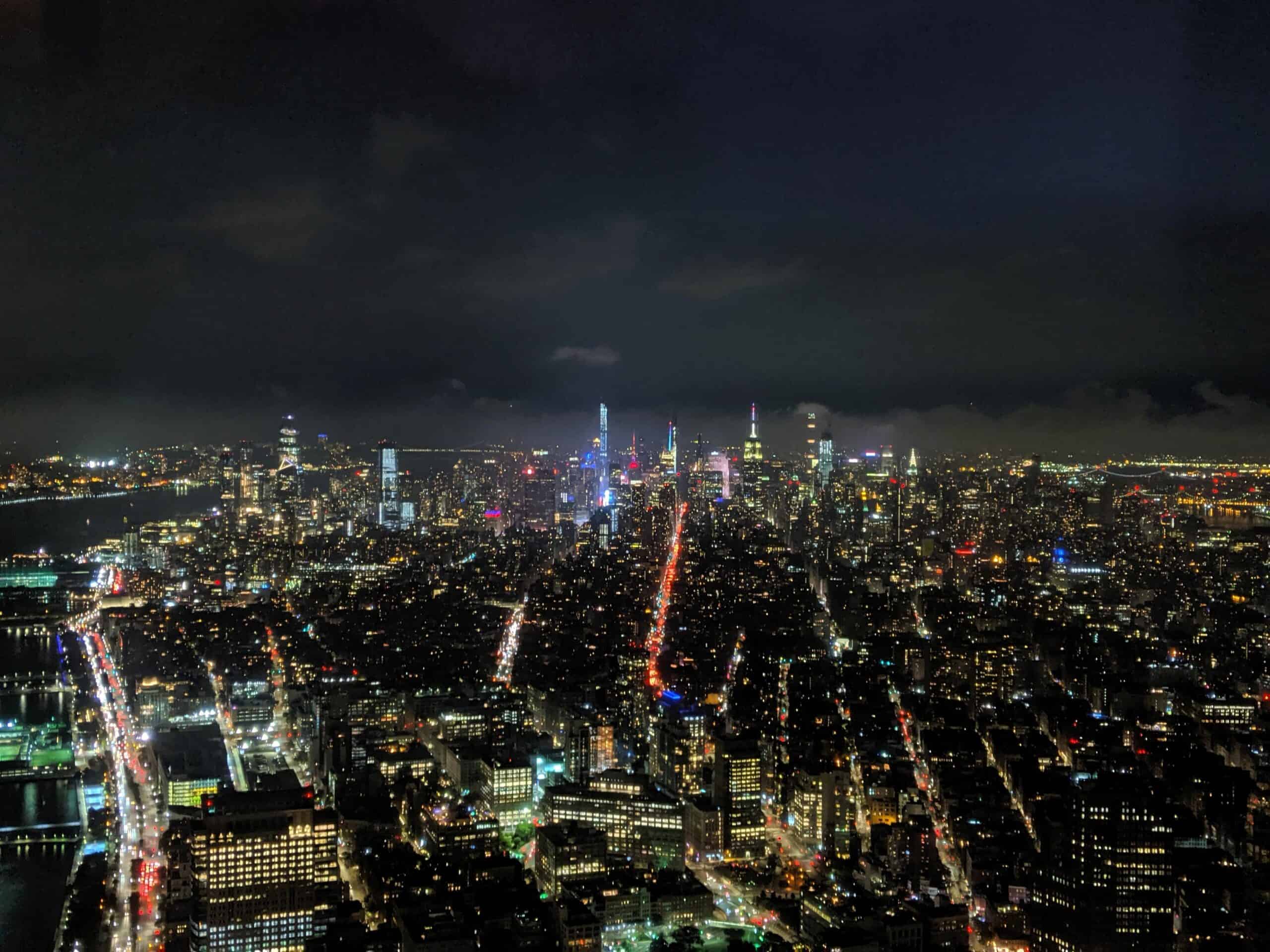 A night shot from One World Trade captures the glittering expanse of Manhattan, with towering skyscrapers and a grid of illuminated streets stretching into the distance. The Empire State Building is visible among the distant high-rises. The Hudson River, lined with more lights, is visible to the left, while the distant horizon is a blur of city glow.