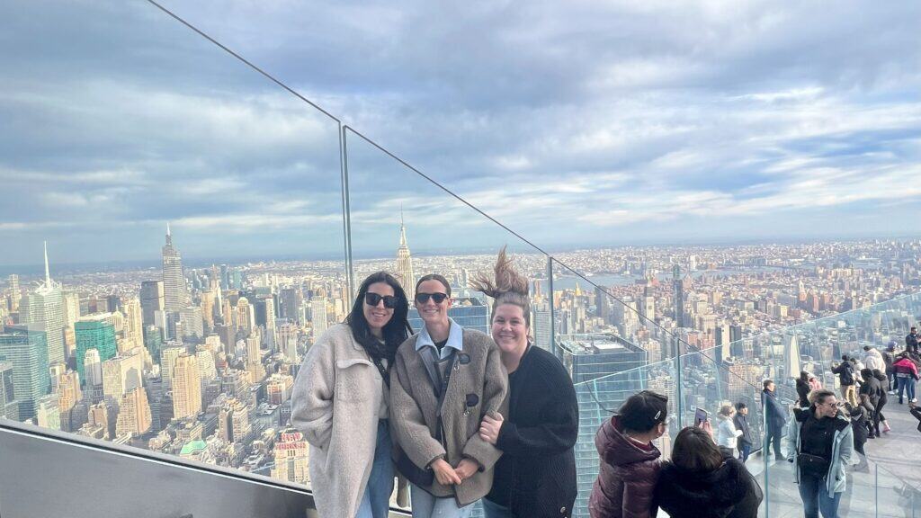 Three women smiling and posing together at The Edge observation deck in New York City, with the Empire State Building and Midtown Manhattan skyline visible in the background under a partly cloudy sky. Other visitors are scattered around the deck enjoying the view.