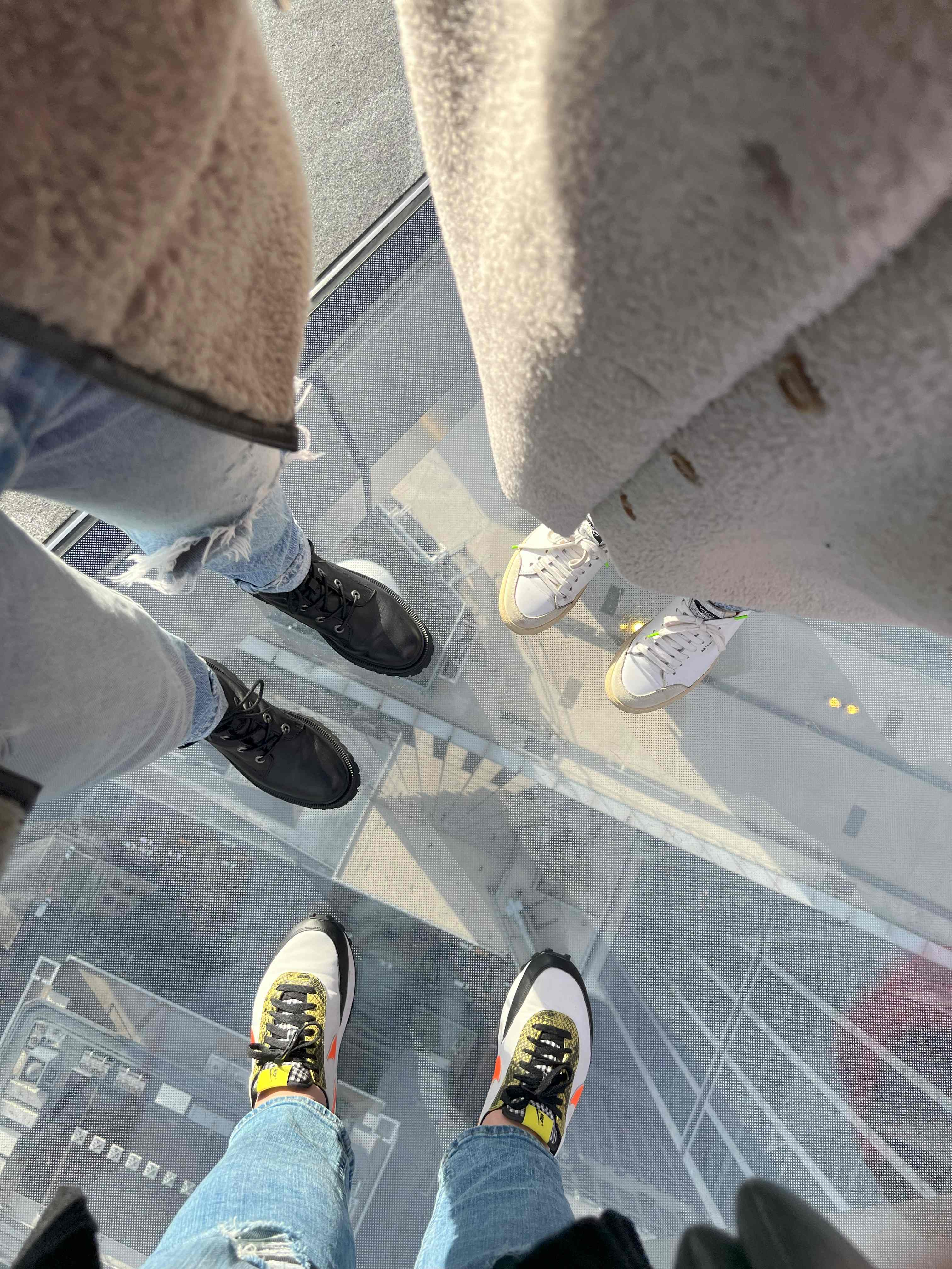 Three people standing on the glass floor at The Edge observation deck in New York City, looking straight down at streets and buildings far below.