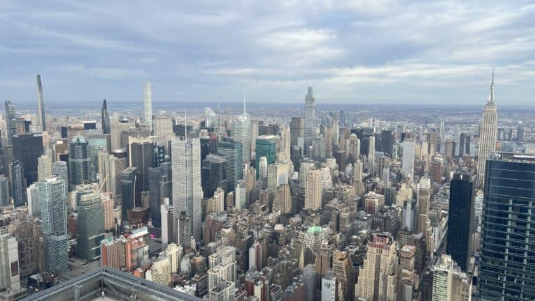 Panoramic view of the Midtown Manhattan skyline from The Edge observation deck, featuring the Empire State Building on the right, modern skyscrapers like One Vanderbilt and 432 Park Avenue, and a mix of high-rises stretching toward the horizon under a partly cloudy sky.