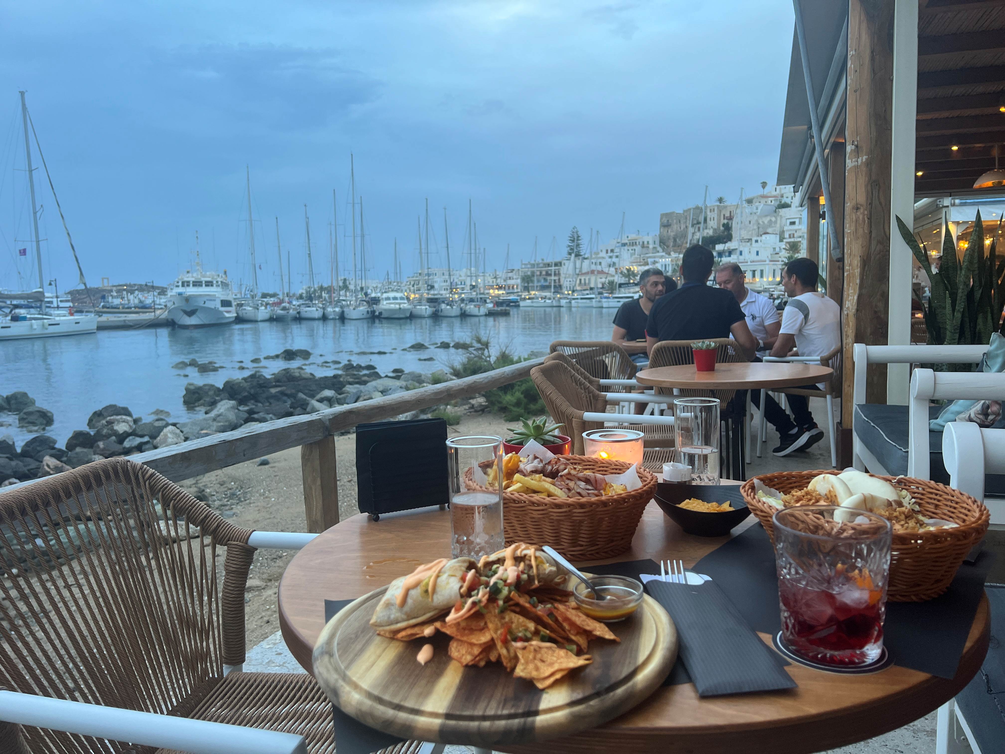 A waterfront restaurant table in Naxos Town, Greece, overlooking the harbor at dusk. The table features grilled seafood with pita bread, bread baskets, and drinks, with sailboats moored in the marina and traditional white Cycladic buildings rising up the hillside in the background