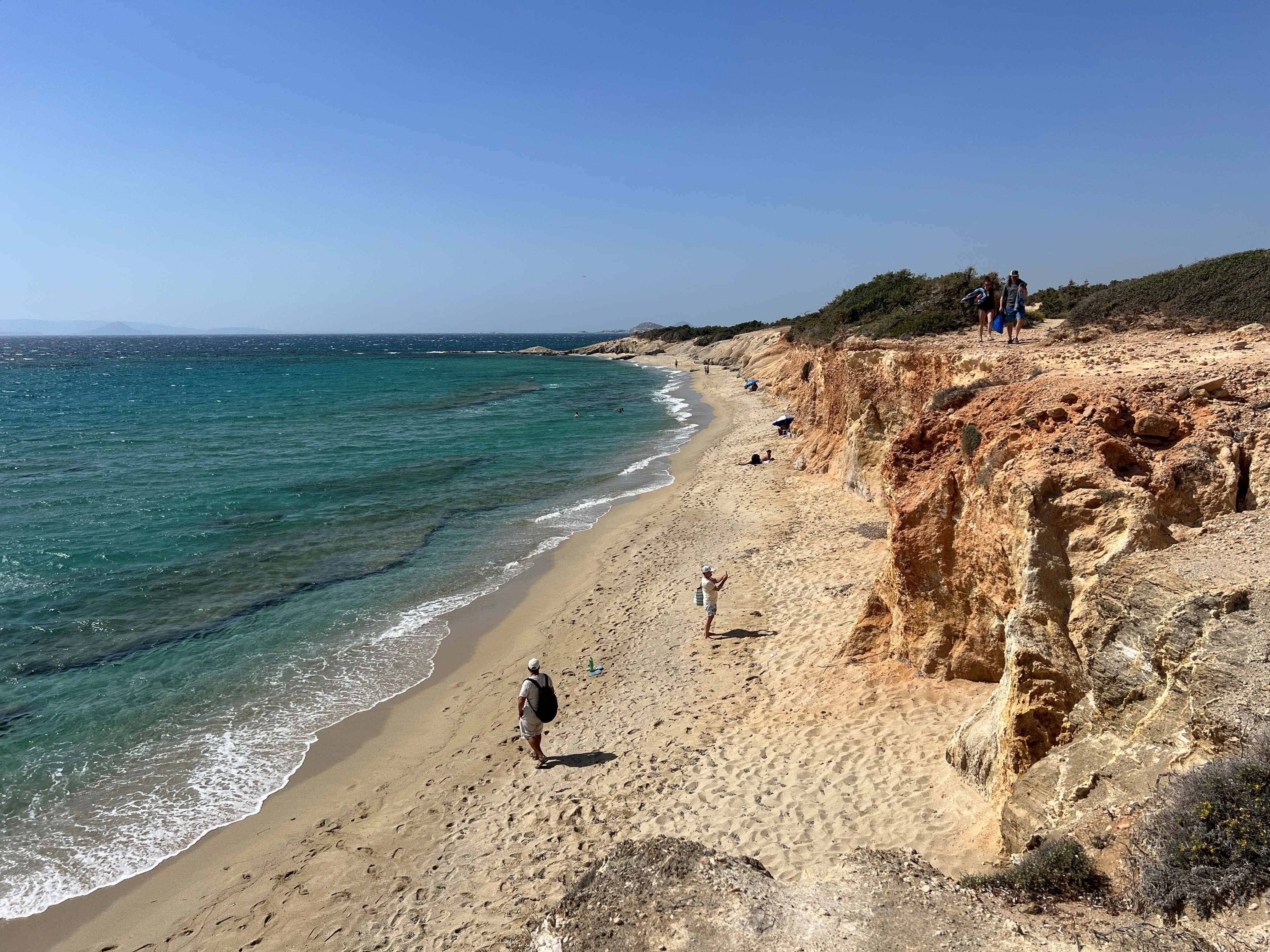 Alyko Beach in Naxos, Greece, featuring a pristine sandy coastline with turquoise waters and dramatic golden limestone cliffs. Several beachgoers walk along the shore and relax on the sand, while green vegetation covers the clifftops under a clear blue sky.