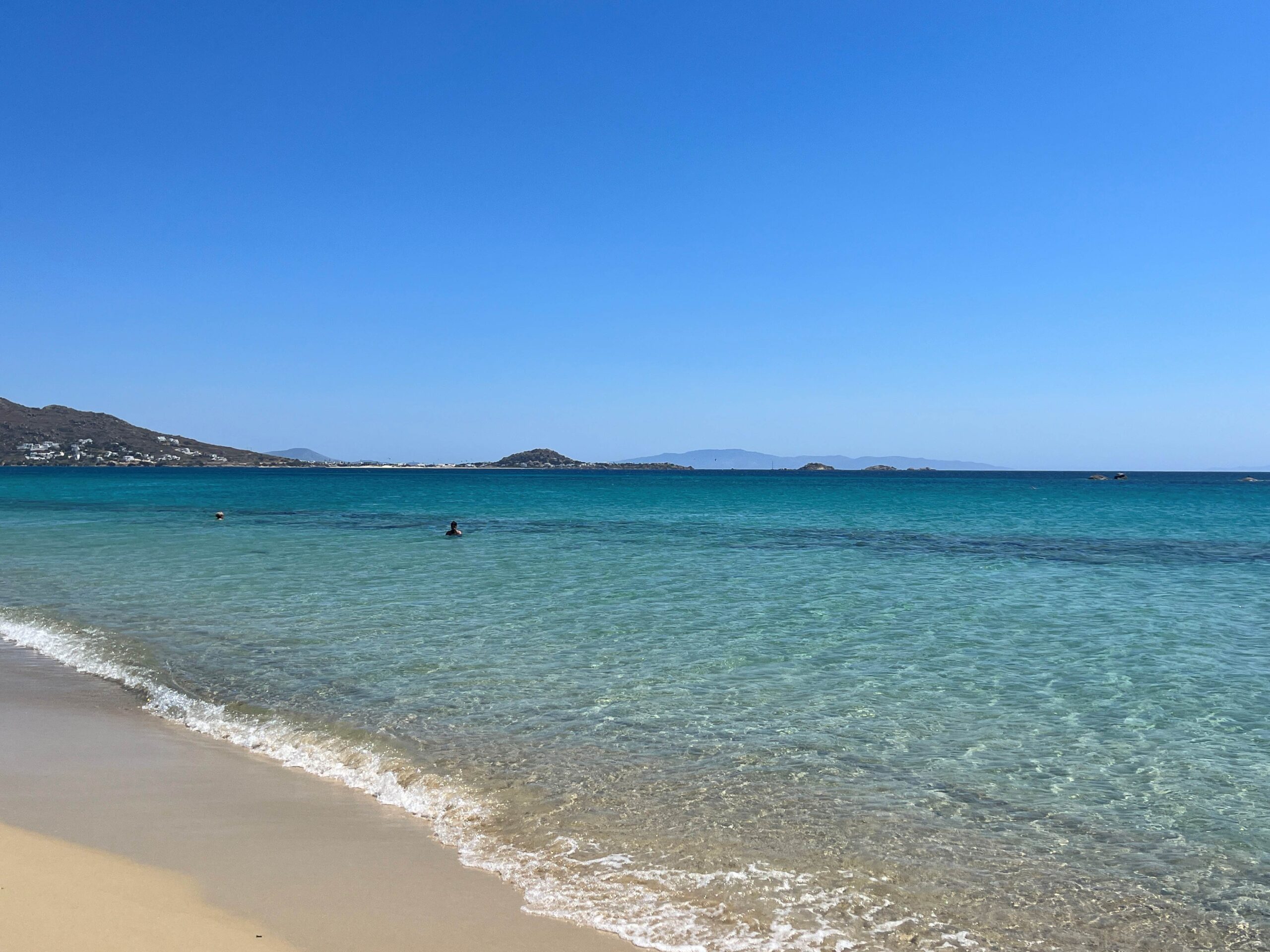 A beach on the island of Naxos, Greece. The foreground shows a smooth, light-colored sandy beach and gentle waves of transparent water rolling onto the shore. The sea extends into a light turquoise color and then a deep blue, with small, rocky islands visible in the distance. The background features a large hill with white buildings on its slopes, and a clear blue sky overhead.