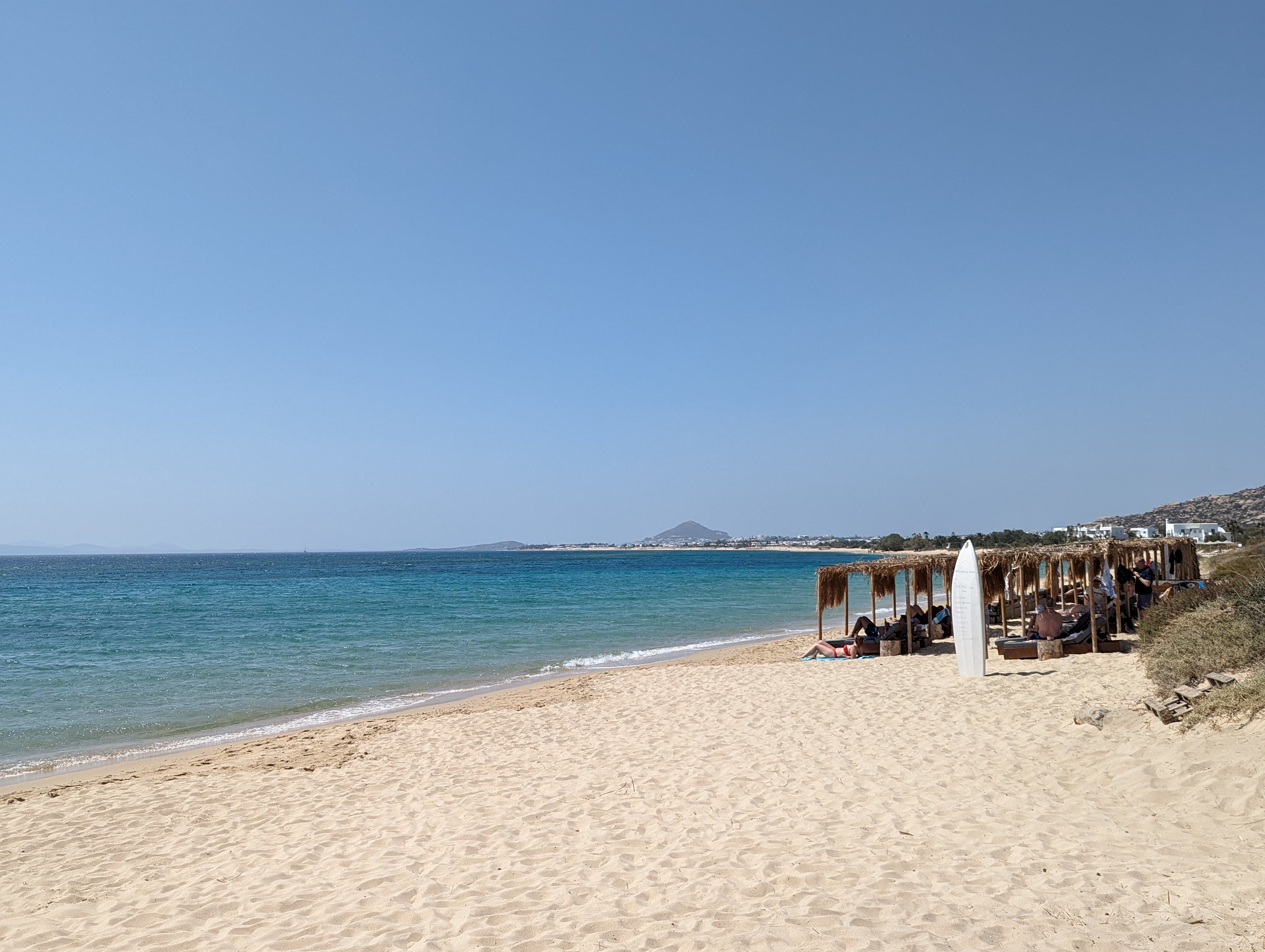 Plaka Beach on the island of Naxos, Greece. The foreground features a wide expanse of light-colored sand. To the right, a line of straw-roofed beach huts and sun loungers faces the sea. The water is a brilliant turquoise that fades into a deeper blue on the horizon. A distant hilly landscape is visible across the sea, and the sky is a clear, cloudless blue.