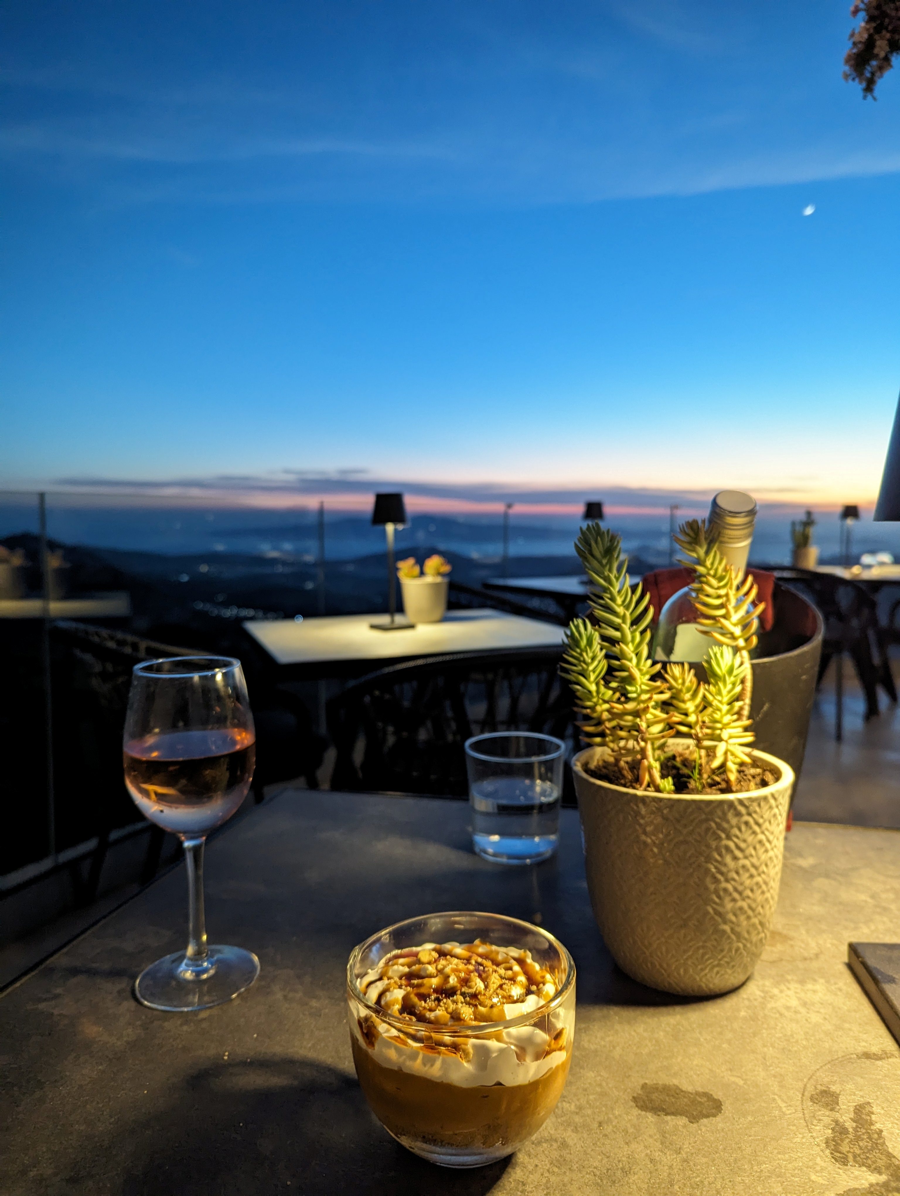 View of a dessert and a glass of wine on a table at an outdoor restaurant. The dessert is a parfait in a glass dish, topped with whipped cream, nuts, and caramel sauce. The table also holds a potted succulent plant and a glass of water. In the background, the sun has set, casting a deep orange and blue hue across the horizon. The view overlooks a sprawling landscape.