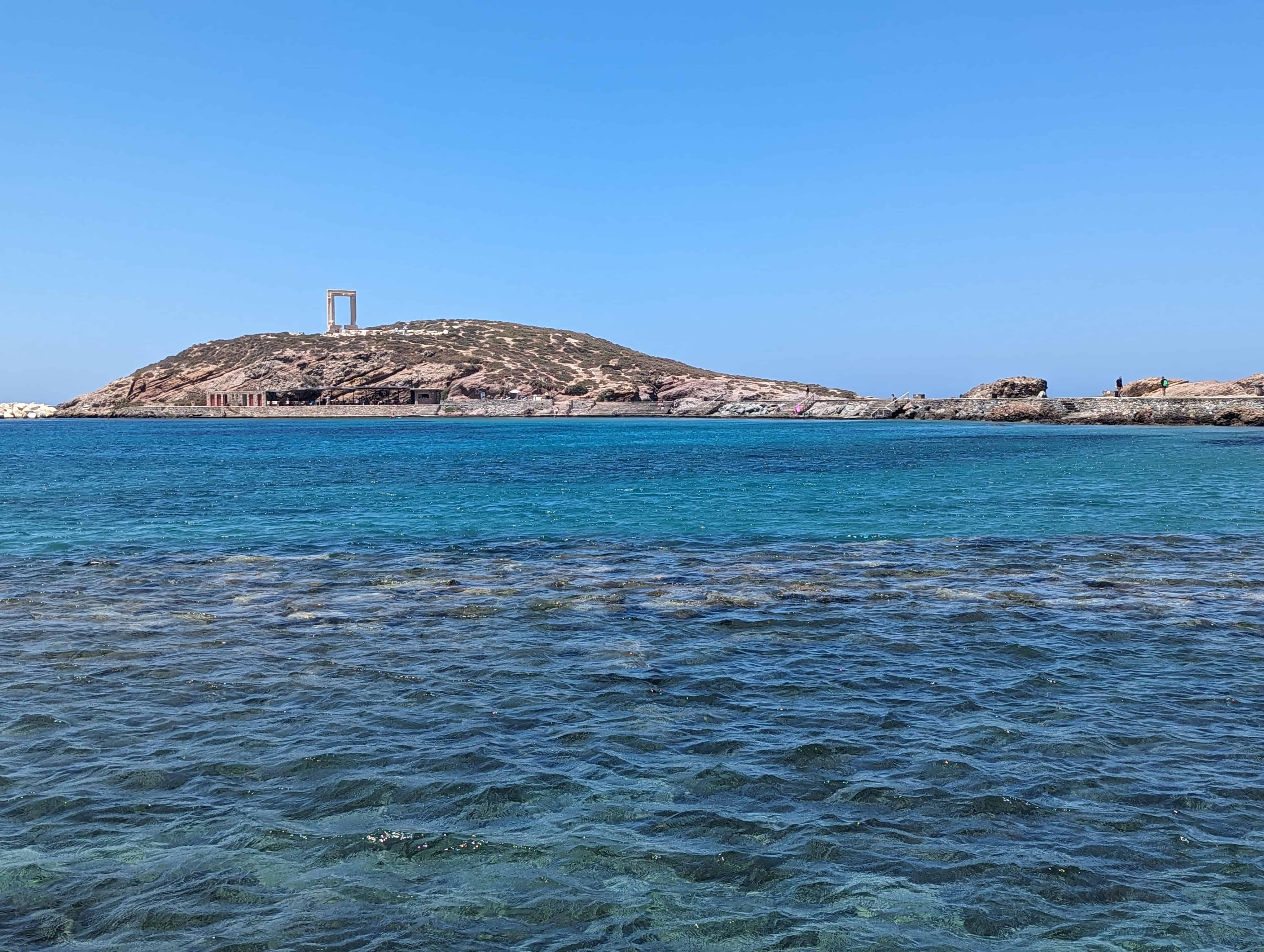 The Portara, a large marble gate, standing on top of a small, rocky hill. The gate is the remains of an ancient temple dedicated to Apollo on the island of Naxos, Greece. The hill is surrounded by the clear blue waters of the Aegean Sea, which is calm and shows some rocky seabed near the foreground. The sky is a cloudless, bright blue