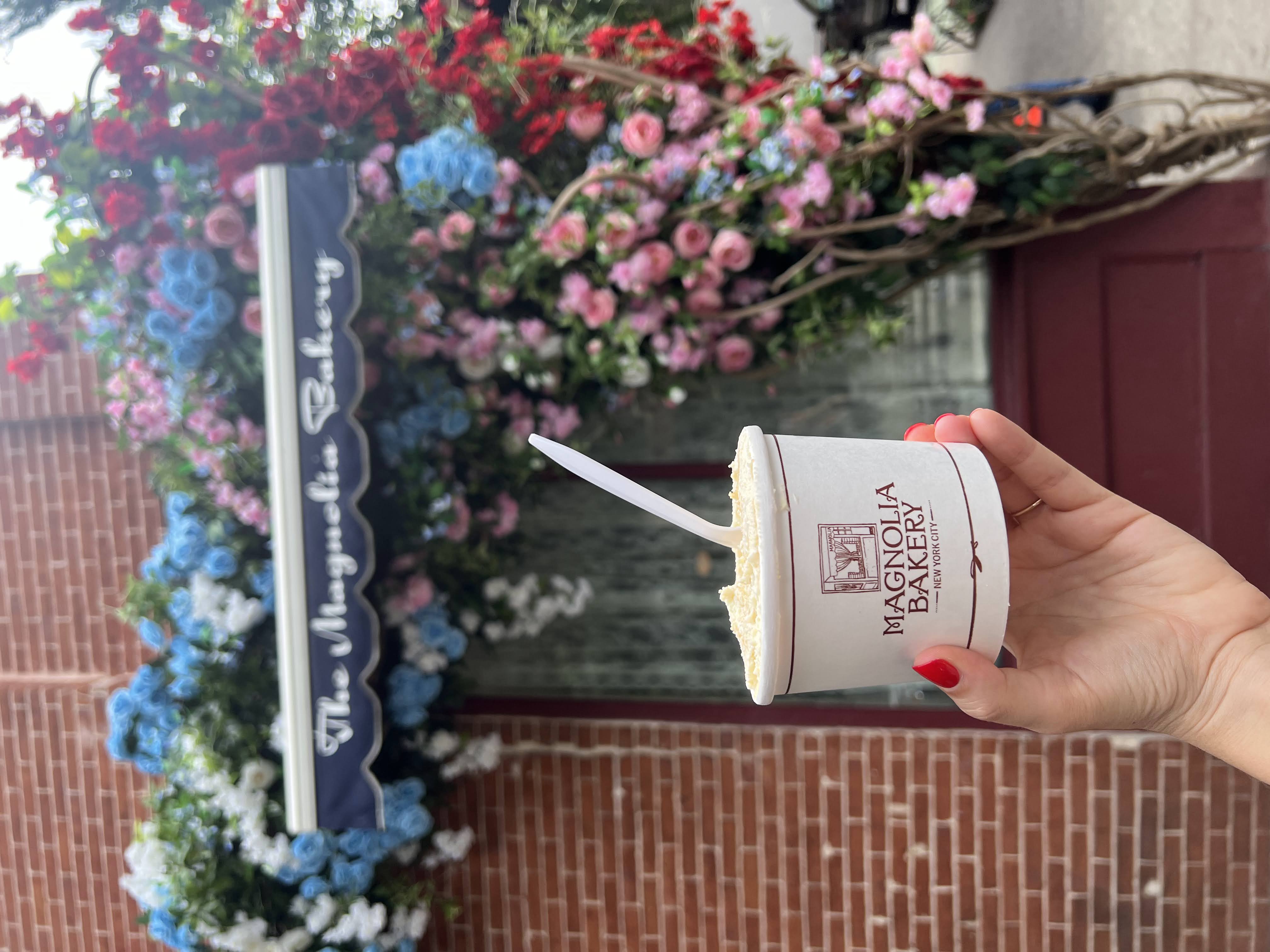 A hand holding a cup of banana pudding from Magnolia Bakery in New York City, with the bakery’s storefront decorated in colorful flowers in the background.