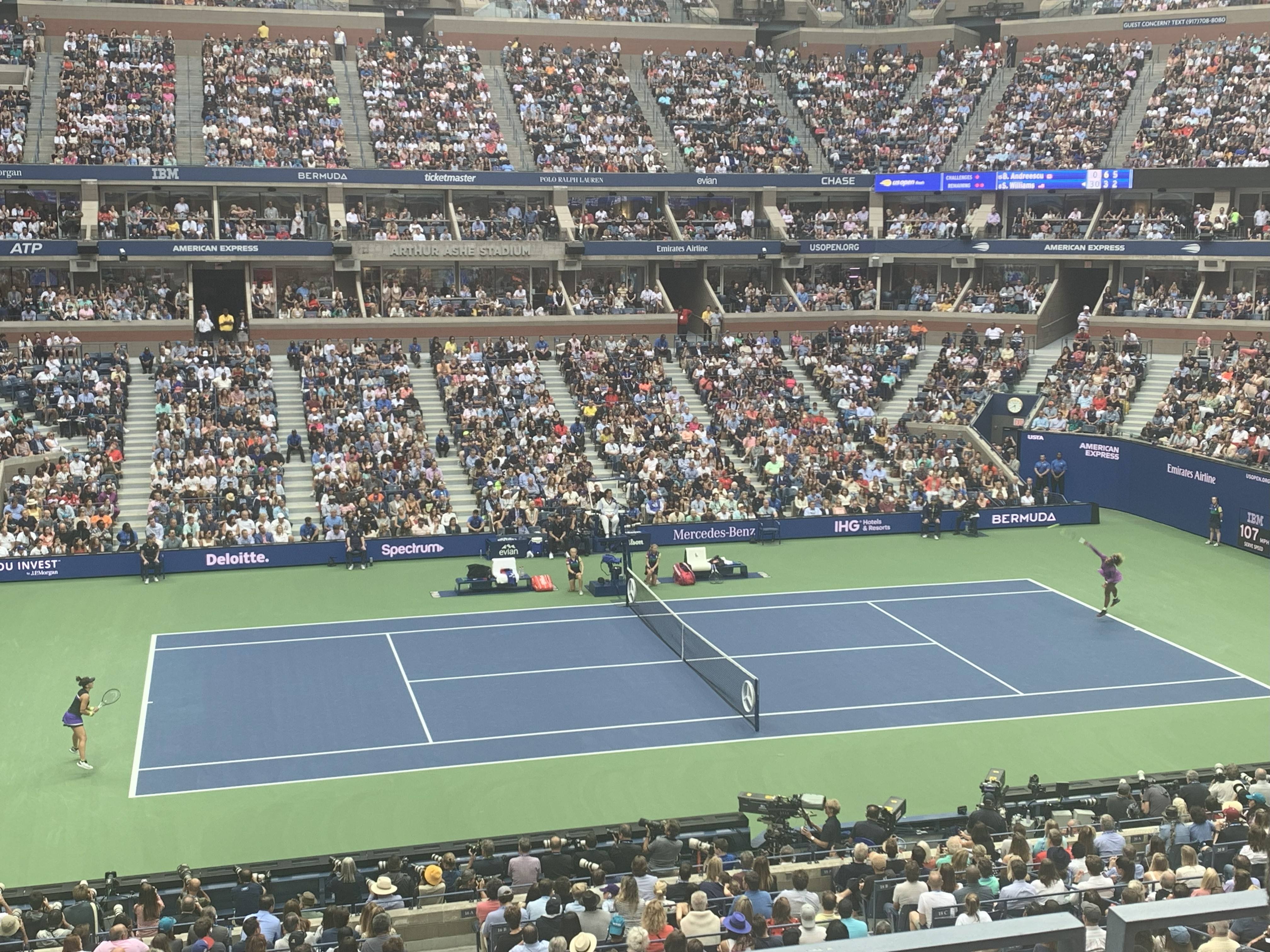 A packed Arthur Ashe Stadium during the US Open, with two tennis players in action on the blue hard court while the crowd watches from the stands.