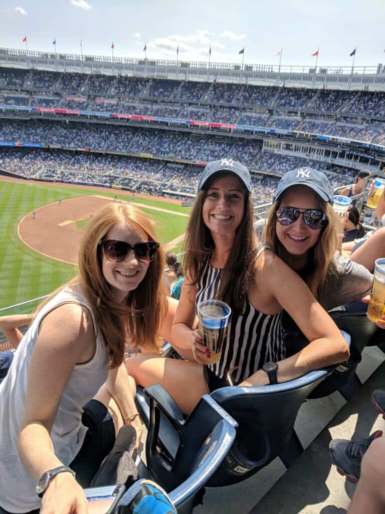 Three smiling women wearing New York Yankees hats and sunglasses, sitting in the stadium stands on a sunny day. They're enjoying drinks, with a packed Yankee Stadium and the green field visible in the background.