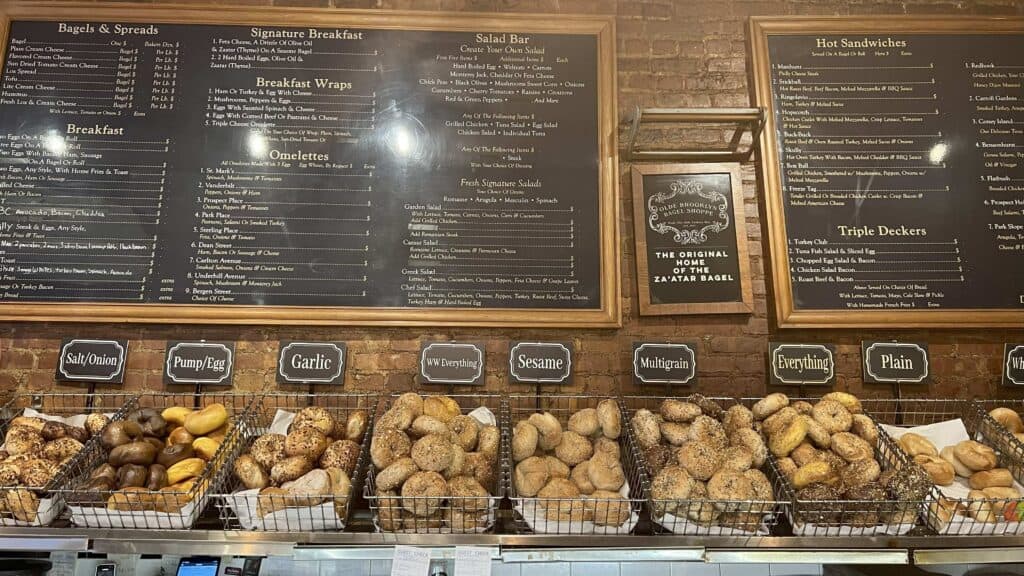 Bagel shop interior with baskets of various bagels (Salt/Onion, Pump/Egg, Garlic, WW Everything, Sesame, Multigrain, Everything, Plain) in the foreground and a large menu board on a brick wall in the background.