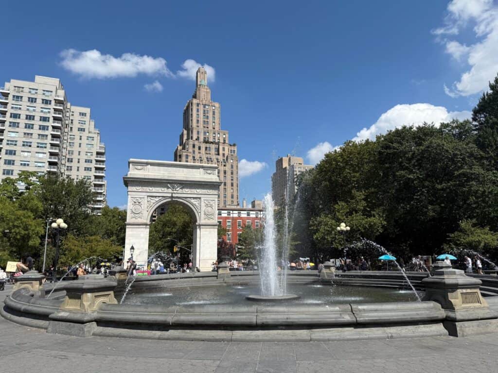 The fountain at Washington Square Park in New York City, with water spraying upward on a sunny day. Behind it stands the iconic Washington Square Arch, framed by leafy green trees and tall city buildings under a bright blue sky with scattered clouds.