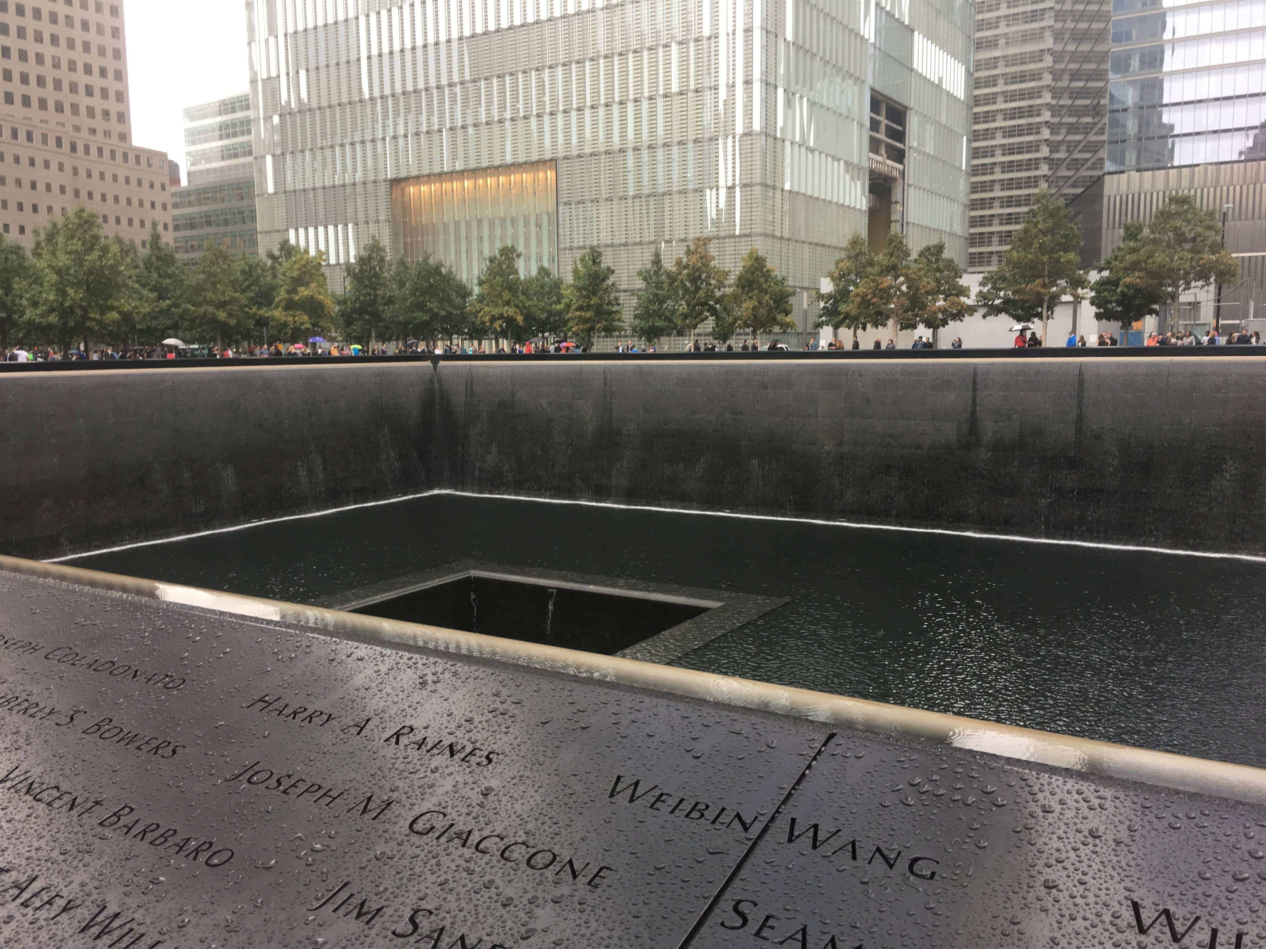 The 9/11 Memorial at the World Trade Center site in New York City, showing one of the large square reflecting pools with water cascading down the walls. Names of victims are inscribed on the bronze panels surrounding the pool, with raindrops visible on the surface. Modern glass buildings and a line of trees stand in the background.
