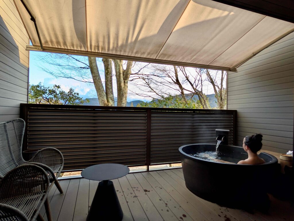 Private outdoor onsen bath at Nol Hakone Hotel with a deep soaking tub on a covered balcony, overlooking trees and mountain views, with a seating area and soft natural light creating a serene atmosphere.