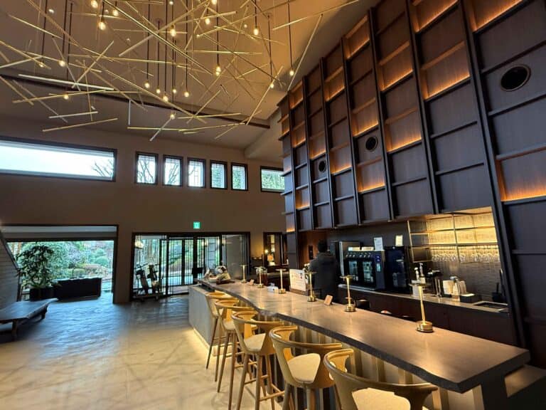Modern, minimalist lounge at Nol Hakone Hotel featuring a long stone bar with wooden stools, warm ambient lighting, and a striking ceiling installation of hanging geometric light fixtures, with large windows overlooking a serene garden.