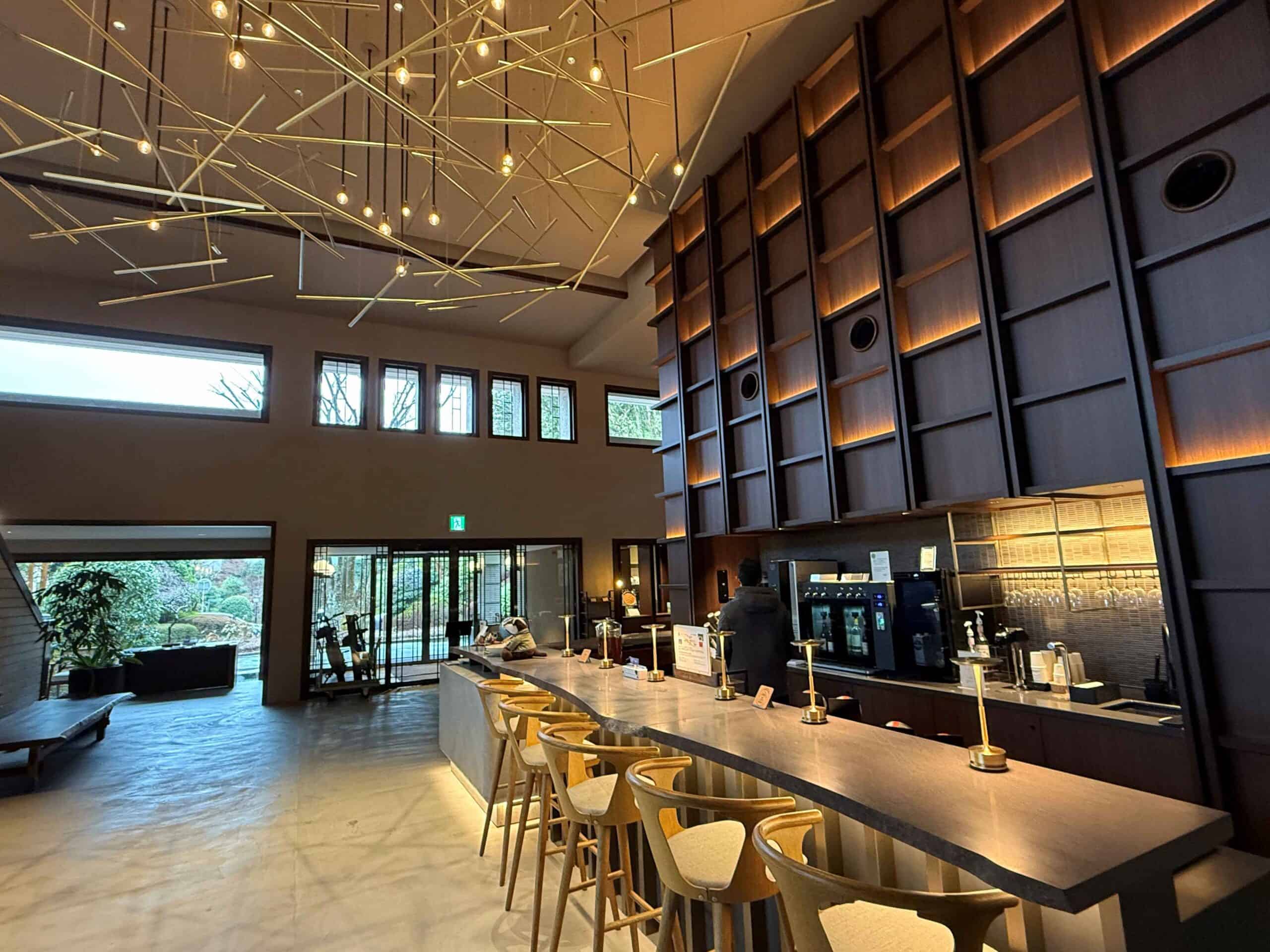 Modern, minimalist lounge at Nol Hakone Hotel featuring a long stone bar with wooden stools, warm ambient lighting, and a striking ceiling installation of hanging geometric light fixtures, with large windows overlooking a serene garden.