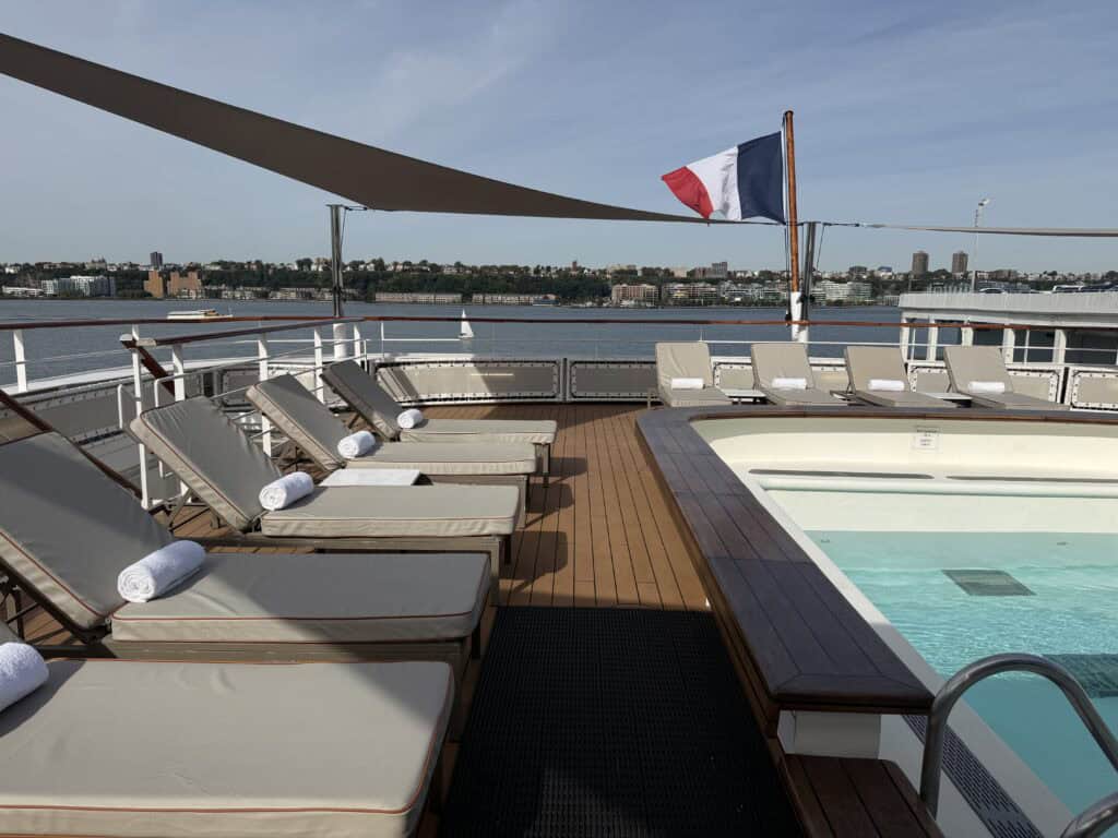 Outdoor pool aboard Ponant’s L’Austral, surrounded by cushioned sun loungers beneath shade sails, with a French flag and water views.