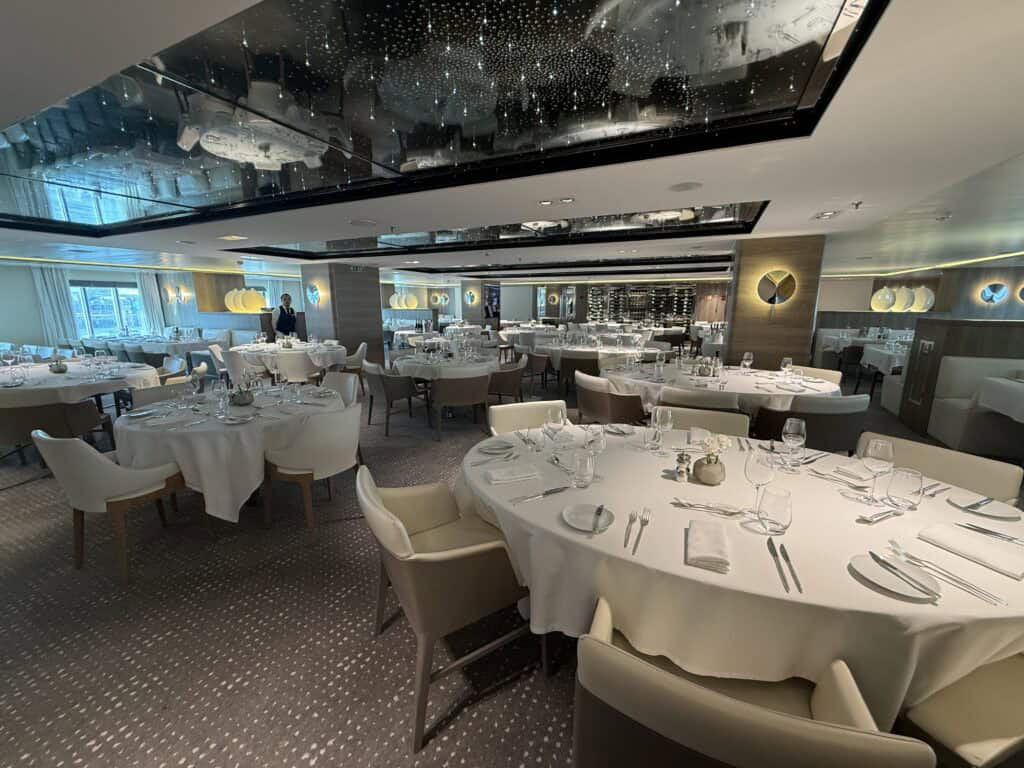 Dining room aboard Ponant’s L’Austral, with white tablecloths, formal place settings, cream chairs and decorative mirrored ceiling panels.